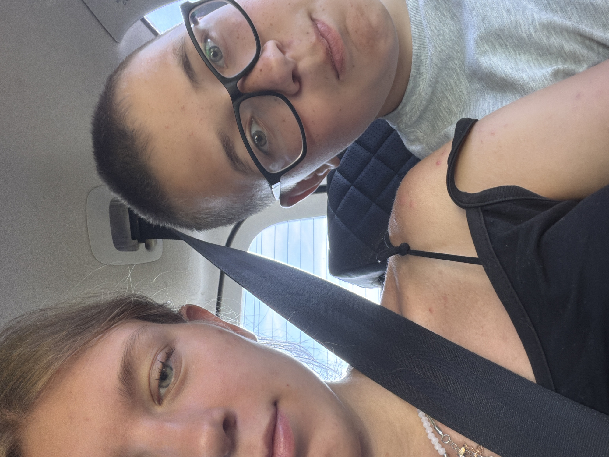 This is a close-up selfie of two young people, likely siblings, in a car. The person on the left, a female, has fair skin and light brown hair pulled back. She is wearing a black tank top and a necklace. Her left shoulder and upper chest are visible, with a seatbelt across her. The person on the right, a male, has fair skin, short brown hair, and is wearing glasses and a grey t-shirt. His face is angled towards the camera. The background shows the interior of a car, including a headrest and a window with a view of trees and the sky. The lighting suggests it is daytime. The image does not contain any information or context related to the city of Kruglovka, Russia.