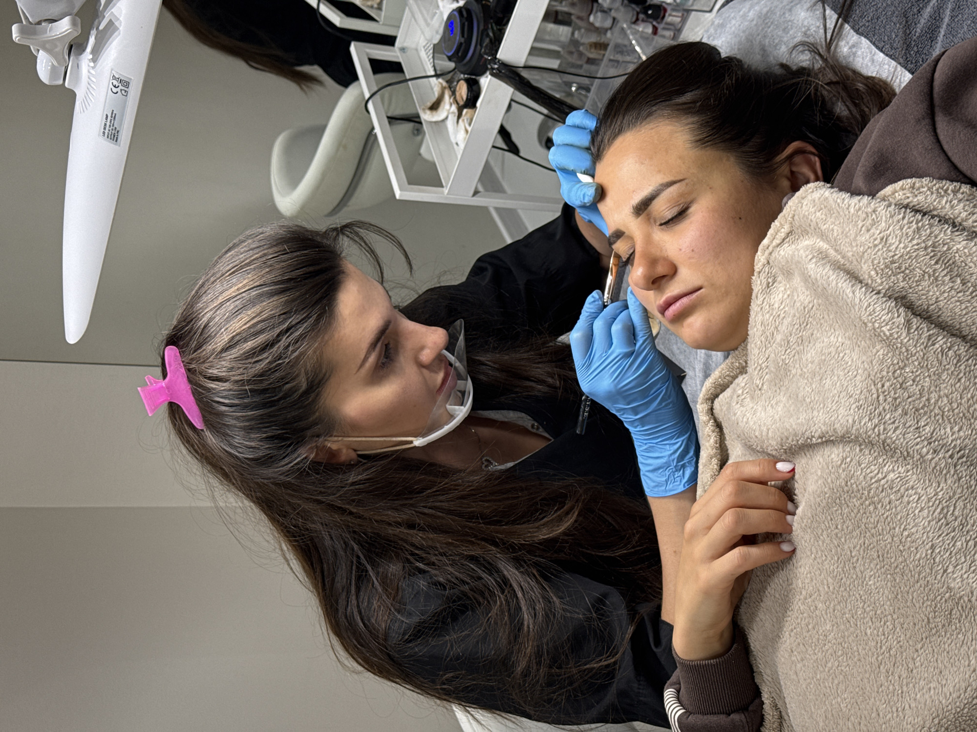 In an indoor setting, a woman with long, dark hair and a pink clip in her hair applies something to the eyebrow of a woman lying down with her eyes closed. The woman applying the substance is wearing blue gloves and a clear plastic face shield. The woman receiving the treatment is wrapped in a soft, beige blanket and appears relaxed. A lamp and various containers are visible in the background. It cannot be confirmed if this scene is in Budva, Montenegro.
