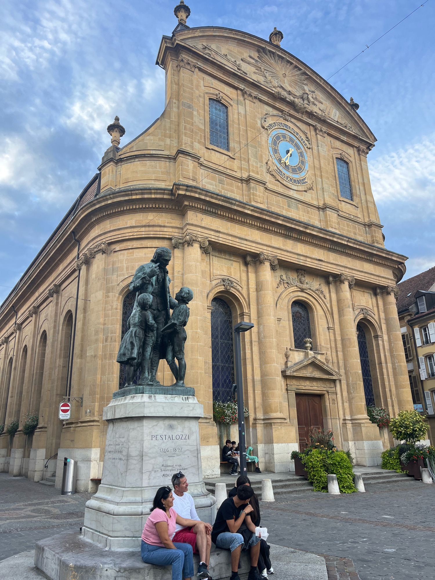 The image depicts the exterior of the Church of Notre-Dame in Yverdon-les-Bains, Switzerland. In the foreground, a bronze statue of a man with three children stands on a marble pedestal. The inscription on the pedestal reads "PESTALOZZI 1746-1827". A group of four individuals are seated on the steps of the pedestal. The church is constructed of pale yellow stone and features arched windows and a large circular clock face above the entrance. The sky is overcast with scattered clouds.