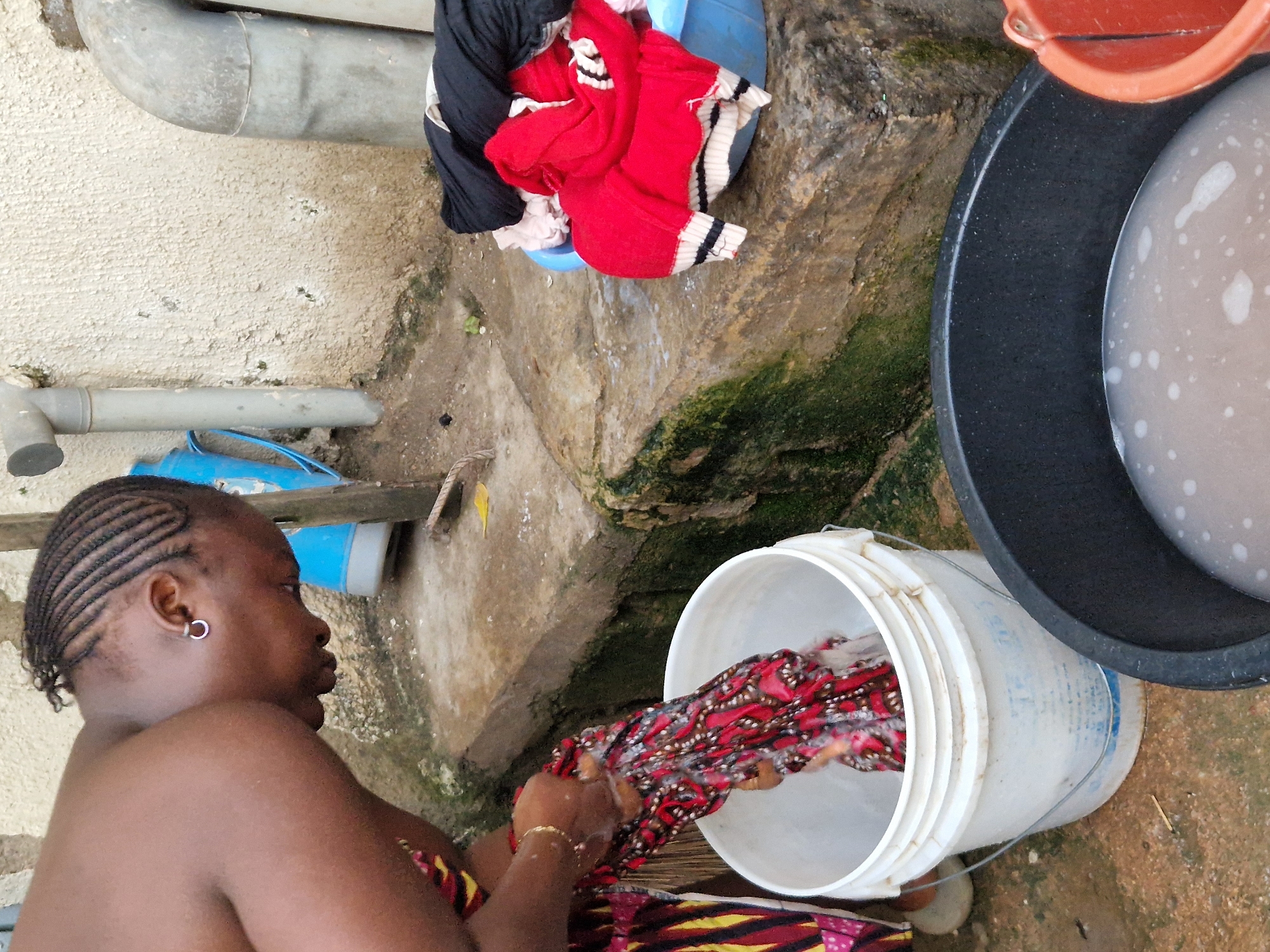 A Black woman, with her hair in cornrows and wearing a silver hoop earring, is shown from the chest up, bent over a white plastic bucket. She is washing a red and white patterned piece of fabric in the bucket. Her hands are wet and soapy. She is wearing a colorful patterned garment on her lower body. To her right, a blue basin is filled with red and black laundry. In the foreground, a large black basin contains soapy water with bubbles. Behind her, a textured cream-colored wall is visible, with gray pipes attached to it. The ground appears to be dirt and stone, with some green moss on the stone. The scene is outdoors, likely in a courtyard or alleyway, in Jos, Nigeria.