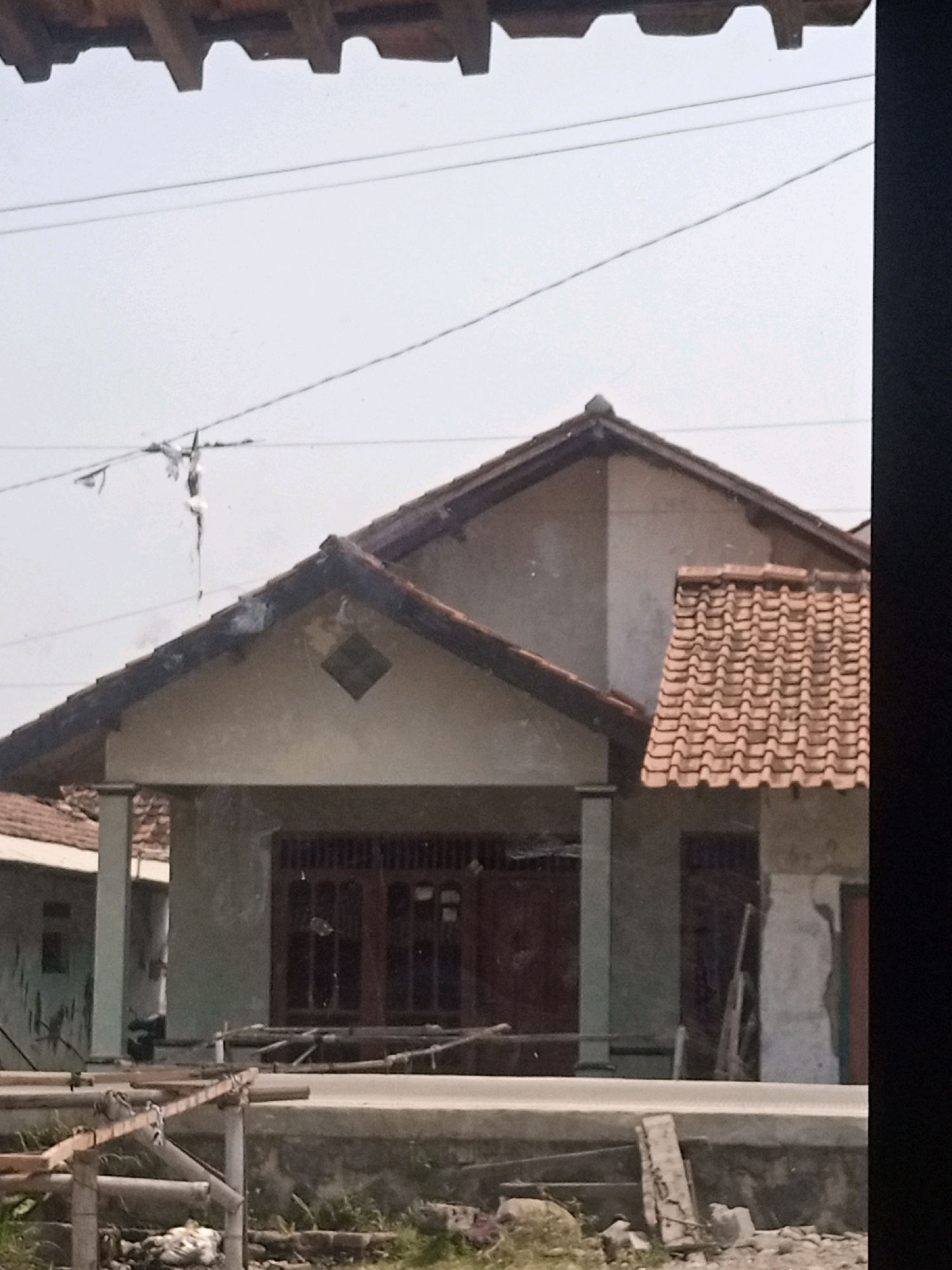 The image shows the exterior of a house in Boyoteluk, Indonesia. The house has a light-colored facade and a tiled roof. The windows are dark brown, and there is a diamond-shaped decorative element on the front of the house. In front of the house, there is a wooden structure that appears to be scaffolding or some form of construction in progress. The sky is overcast.