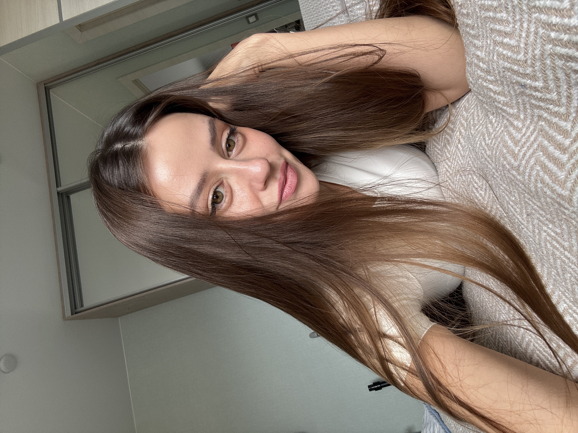 A young woman with long brown hair and fair skin is lying on a bed or couch covered with a beige herringbone-patterned fabric. She is wearing a white top. Her head is positioned towards the upper left of the frame. The background includes a light gray wall and a sliding door closet with a light-colored frame. A white wall outlet is visible on the lower left. There is no definitive visual element to confirm the location as Krasnoyarsk, Russia.