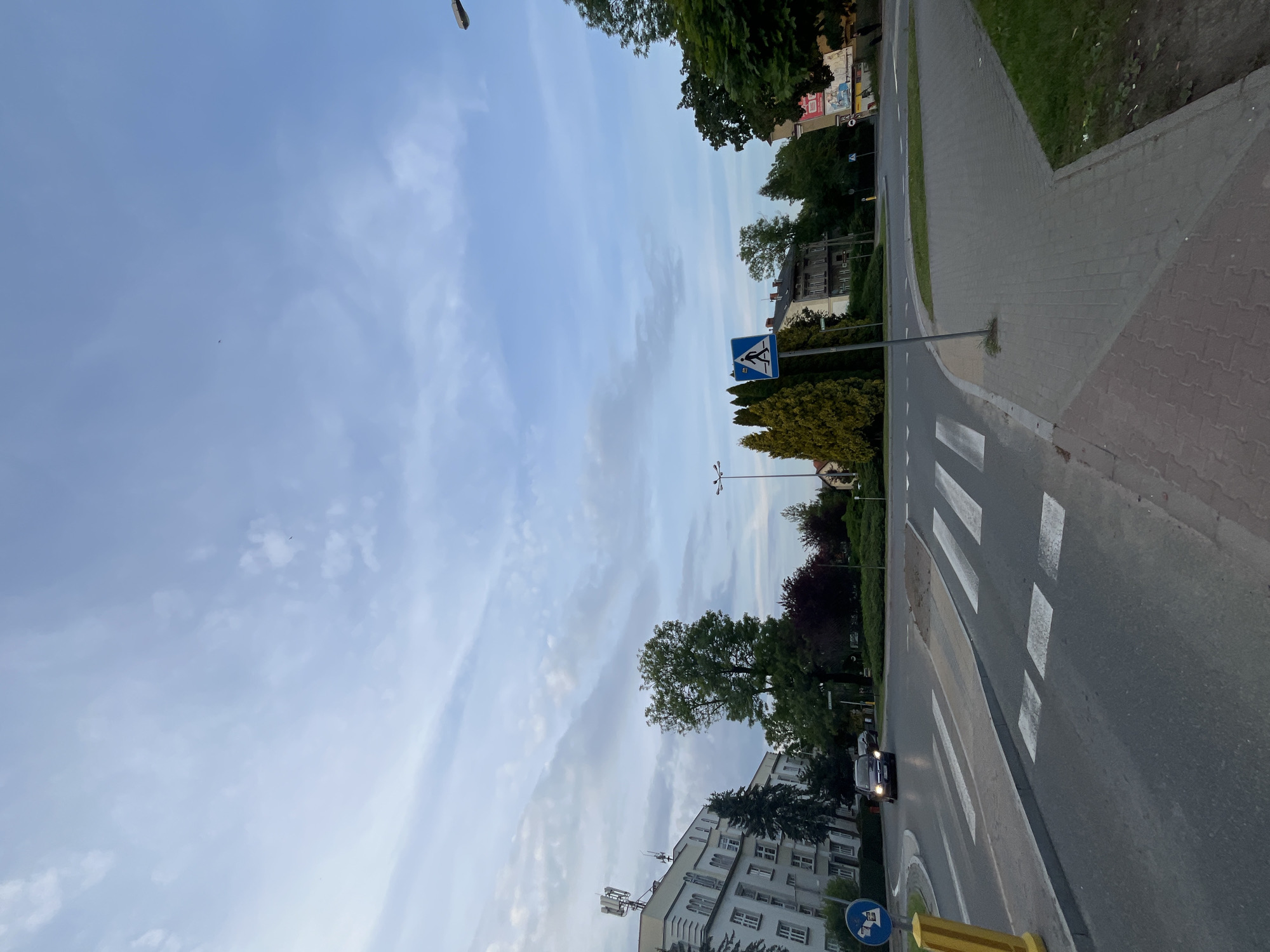 The image depicts a street scene, likely in an urban or suburban setting, captured during late afternoon or early evening twilight. The original photo is rotated 90 degrees counter-clockwise; when viewed upright, the road stretches from the foreground towards the background, gently curving to the right.
Prominently featured is a pedestrian crossing, marked by white zebra stripes across the asphalt road. A dark-colored car with its headlights on is visible on the left side of the road, approaching the crossing. Sidewalks line both sides of the street, with a paved path running parallel to the road on the right, bordered by grass and mature trees. More trees and greenery are scattered throughout the scene, including lush foliage on the right and left sides.
Buildings are visible in the background and on the left. On the left, a multi-story building with numerous windows and several rooftop antennas stands beside the road. In the mid-ground on the right, another building can be seen partially obscured by trees.
A blue square sign with a white triangle and a walking person symbol, indicating a pedestrian crossing, is positioned on the right side of the road. Another blue circular sign with an unclear symbol (possibly an information or newspaper symbol) is visible near the bottom left, partially obscured. A street light or lamppost extends from the top of the frame. The sky above is a light blue with wispy, scattered clouds, suggesting fair weather. No people are visible, and no discernible text can be read on any signs or buildings due to distance or blur.
