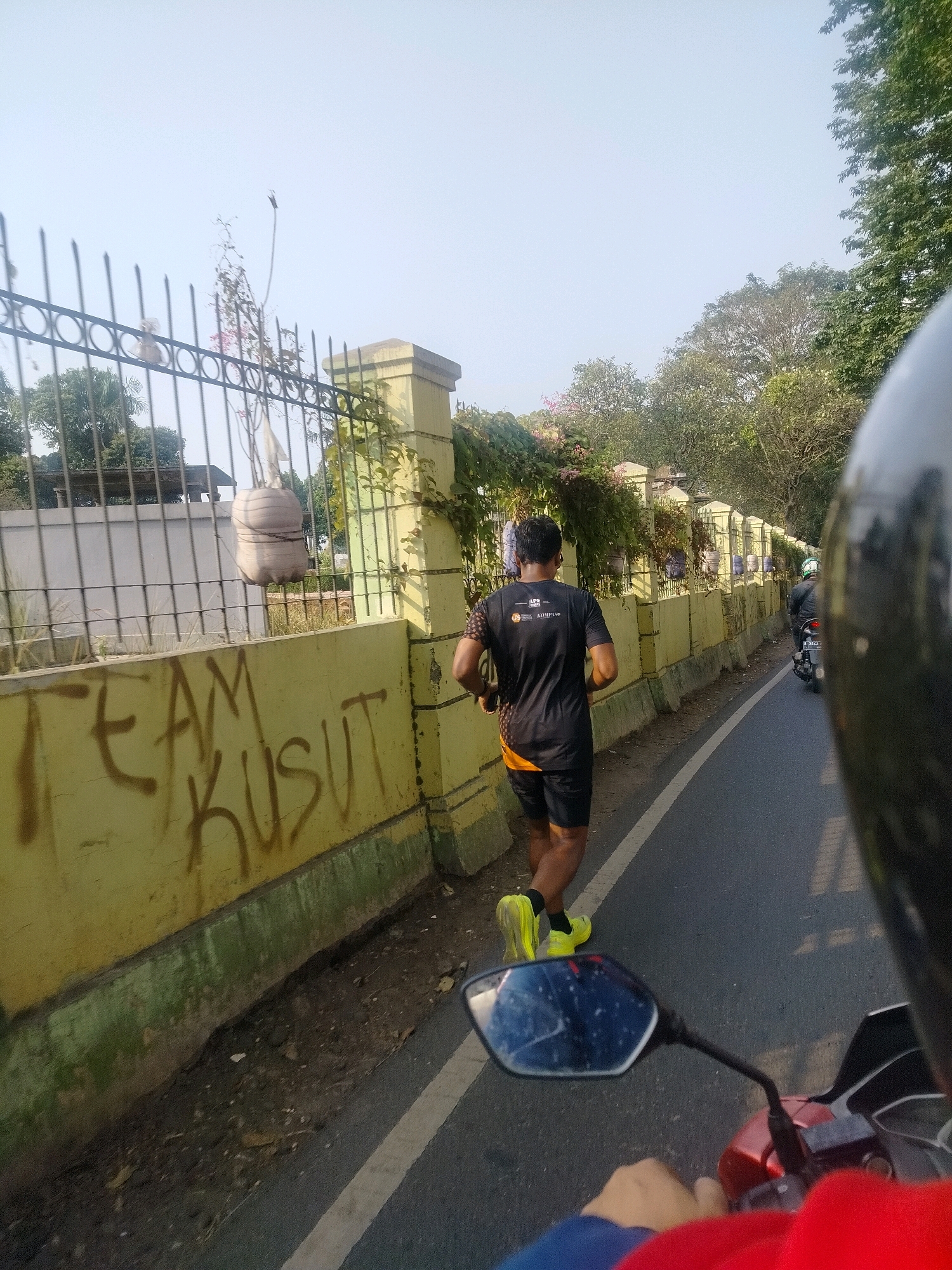 The image captures a man running on a paved road in East Jakarta, Indonesia. The runner, seen from behind, is wearing a black t-shirt with logos and black shorts, and bright yellow running shoes. He is running alongside a low yellow wall, which has graffiti that reads "TEAM KUSUT" in brown paint. The wall is bordered by a strip of dry earth and vegetation. Beyond the wall, a black metal fence runs along the perimeter, with a large potted plant suspended from it. Trees and other foliage are visible in the background, suggesting an outdoor, possibly park-like or residential area. The sky is clear and bright. The perspective of the photo suggests it was taken from a moving vehicle, likely a motorcycle, with the handlebar and part of a helmet visible in the foreground. Another motorcycle with a rider is also visible further down the road.