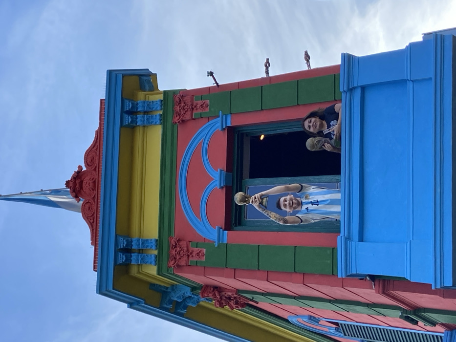 A low-angle shot of a brightly painted building with a blue, white, and red flag waving from the roof. A man in an Argentina jersey stands in a window holding a golden trophy. A woman stands behind him, partially obscured. The building is painted in red, yellow, green, and blue.