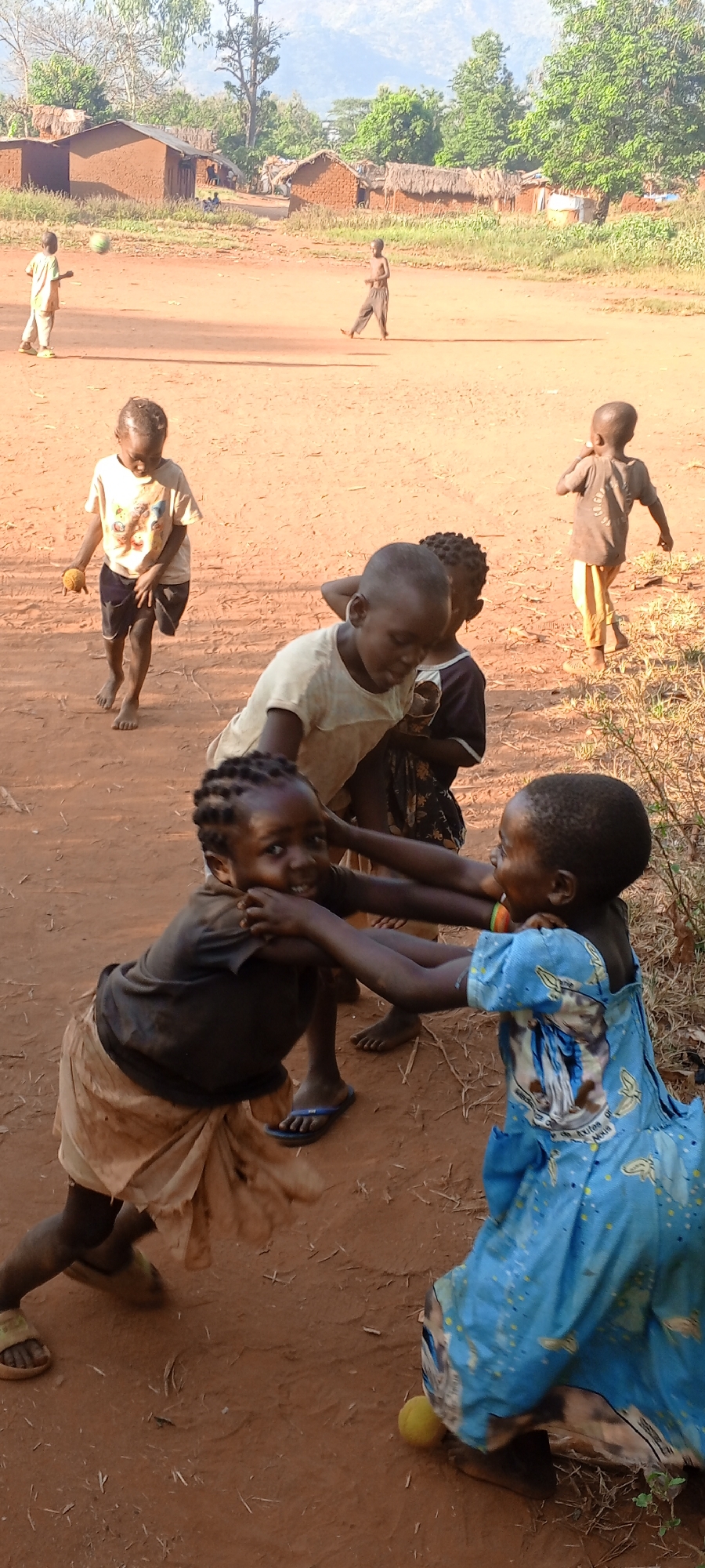 In Ngalula, Democratic Republic of the Congo, a group of children play on a dirt field. Two children in the foreground, one wearing a black shirt and a light-colored skirt and slippers, and the other wearing a blue dress, are engaged in a playful tug-of-war. A yellow ball lies on the ground near the child in the blue dress. Other children are scattered in the background, some walking and others playing with a ball. Houses with thatched roofs can be seen in the distance. Mountains rise in the background behind the village. The ground is reddish-brown dirt.