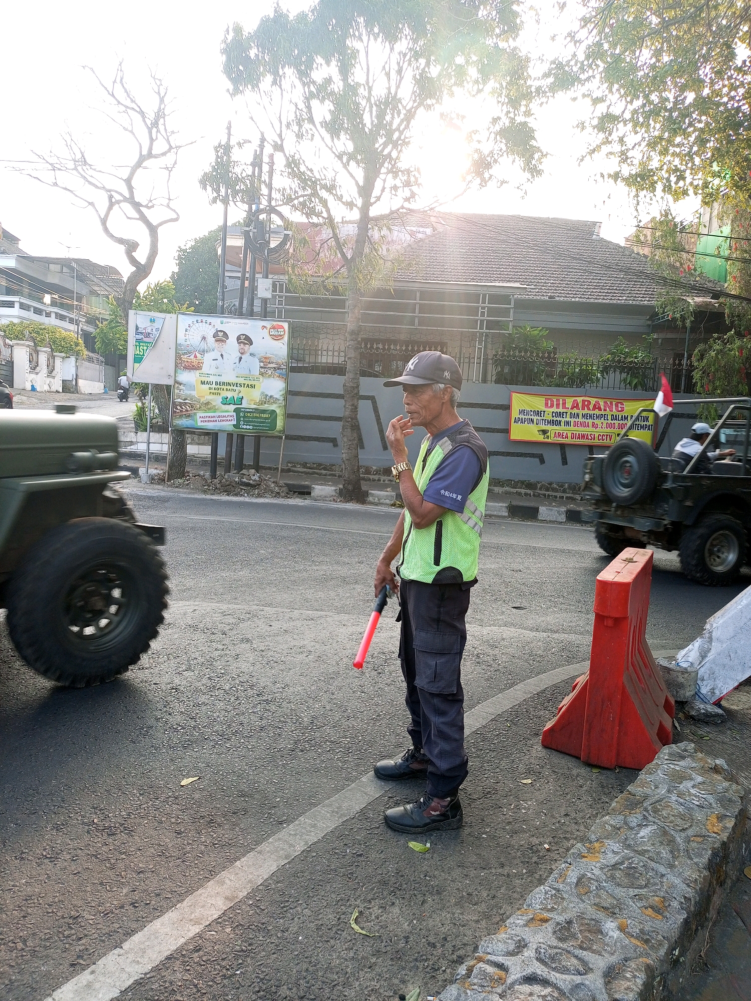 In Batu, Indonesia, on a paved road with a white line, an elderly man wearing a dark baseball cap with a white logo, a dark blue t-shirt, and a bright green reflective vest stands facing right. He is holding a red traffic wand in his right hand, and his left hand is raised to his mouth as if he is smoking or chewing something. He wears dark pants and black shoes. To his left, the large tire of a green military-style vehicle is visible, partially obscuring him. Behind him, a yellow banner with Indonesian text and a red and white flag are attached to a gray wall. A black Jeep with a spare tire on the back drives past him, and an orange traffic barrier is placed on the sidewalk to his right, along with a stone border. The scene is bathed in bright sunlight, with lens flare visible, suggesting it is daytime. Trees with sunlight filtering through their leaves are in the background, along with a house with a tiled roof. A billboard with images of two men in uniforms and text in Indonesian is also visible in the background.