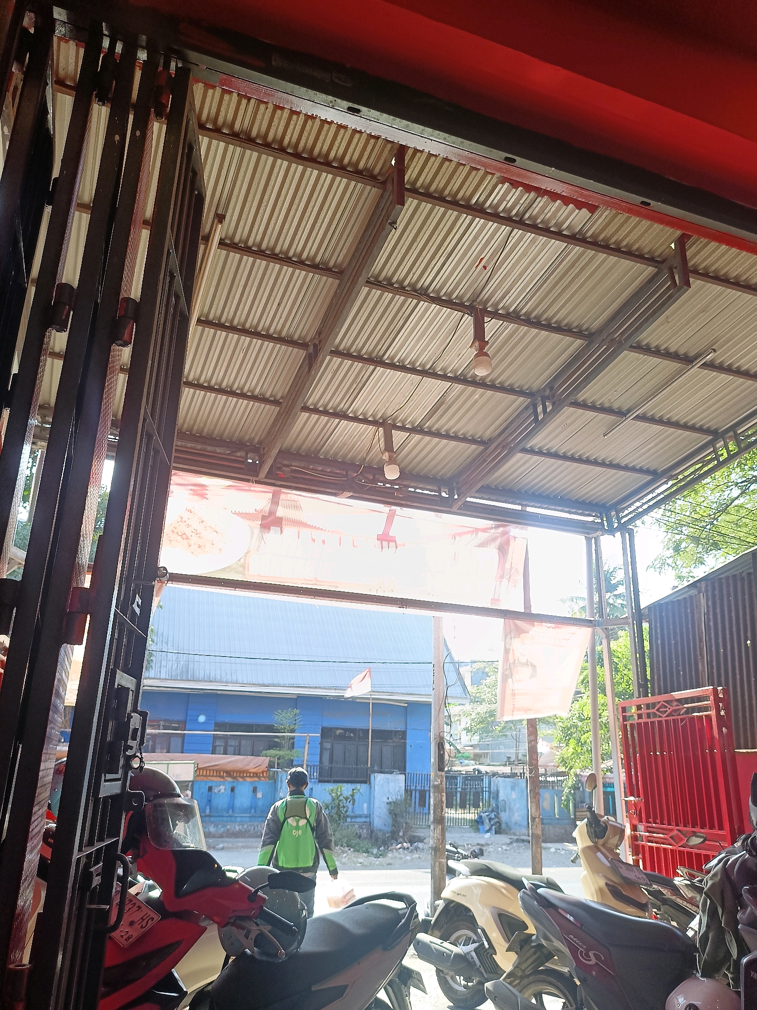 The image captures a view from under a corrugated metal awning, looking out towards a street scene in Makassar, Indonesia. In the foreground, to the left, is a dark metal gate structure. Partially visible are several parked motorcycles. A person wearing a green and gray jacket with a green backpack walks away from the camera, towards the street. Behind the person, there are buildings, one of which is painted blue. A red metal gate is visible to the right of the frame. The awning is supported by metal beams and has lighting fixtures hanging from it. The overall impression is a typical street view in an urban environment.