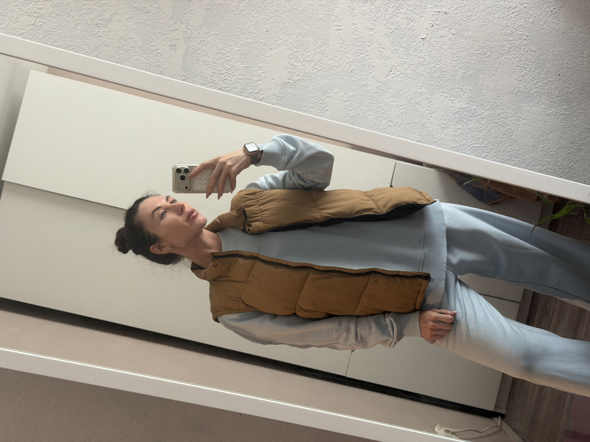 A young woman is captured in a mirror selfie within an indoor setting, identified as Perm, Russia. She is shown from approximately the mid-thighs up, standing with her body angled slightly to her right and her head turned to look towards the left. She holds a light-colored smartphone with a textured case in her right hand, using it to take the picture. Her dark hair is styled in a bun.
She is dressed in a casual outfit consisting of a light blue long-sleeved top, possibly a sweatshirt, and matching light blue sweatpants. Over the top, she wears a brown or mustard-colored puffy vest. A dark-strapped smartwatch is visible on her left wrist.
The background, reflected in the mirror, features a textured white wall above a white horizontal frame, which appears to be the top edge of the mirror. Below this, part of a white cabinet or piece of furniture is visible. A small green plant can be partially seen on the far right side of the reflection. The lighting suggests an indoor daytime scene. Her expression is neutral and contemplative. There is no discernible text visible in the image.