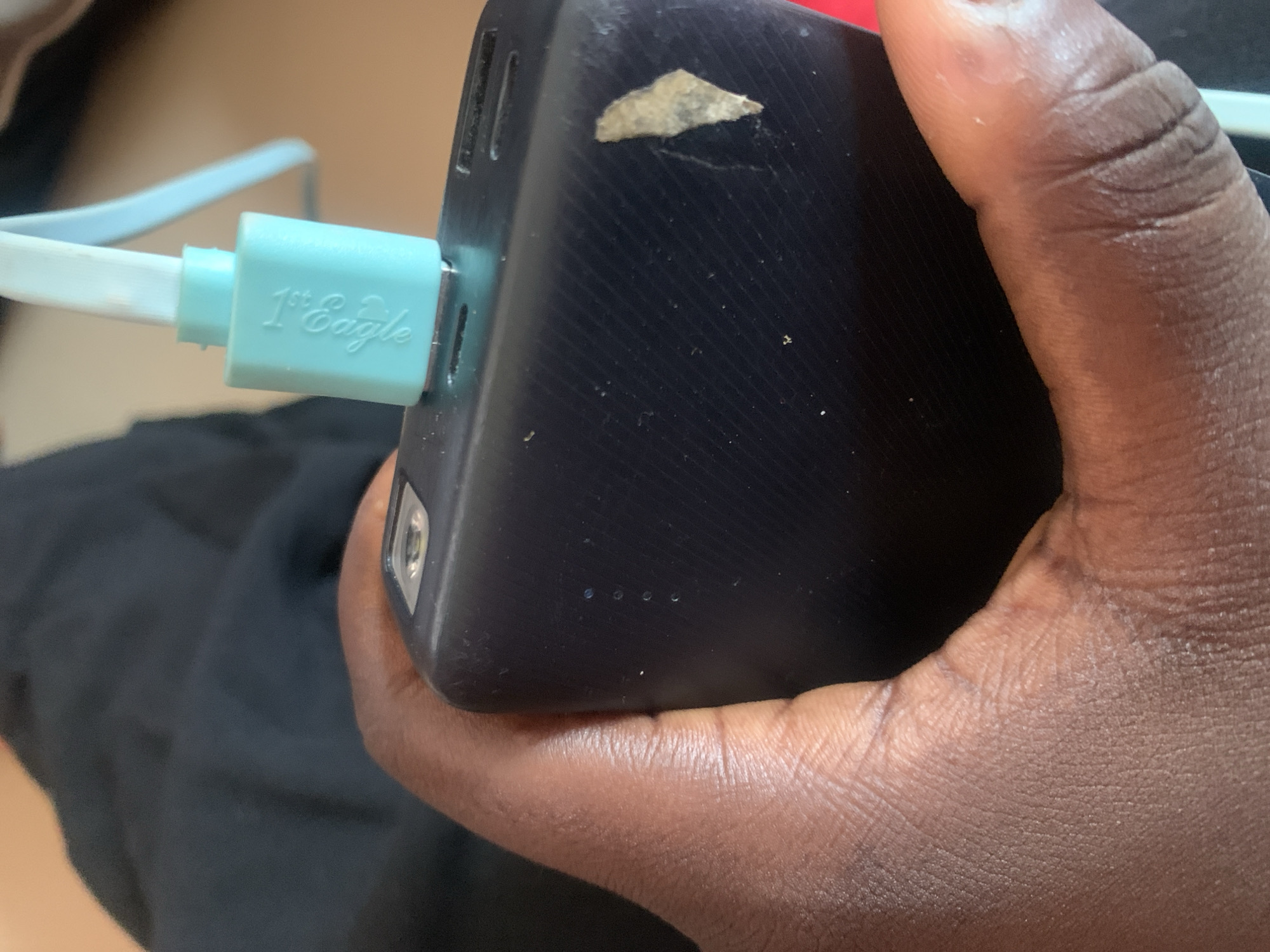 A close-up shot shows a person's hand holding a black power bank. A light blue charging cable is plugged into the power bank, and the connector has "1st Eagle" embossed on it. The background is out of focus, showing dark fabric and a light-colored surface. The scene is indoors, likely a domestic setting in Jauro Yinu, Nigeria.