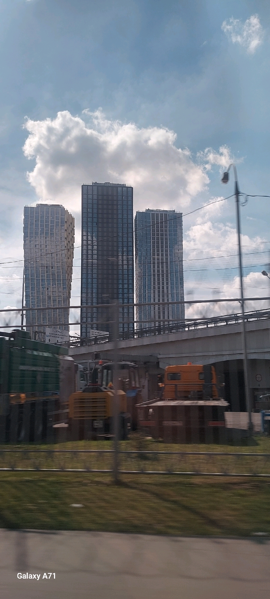 The image depicts a cityscape with three tall, modern high-rise buildings in the background. The buildings are characterized by their grid-like window patterns. In the midground, an elevated overpass or bridge runs horizontally across the frame, with construction vehicles visible beneath it, including a yellow front-end loader and an orange truck. In the foreground, a blurred grassy area with a fence and some pavement suggests the viewpoint is from a moving vehicle. The sky is partly cloudy, with fluffy white clouds against a blue backdrop. A street lamp is visible on the right side of the image. The overall impression is one of urban development and construction, set against a bright, daytime sky.