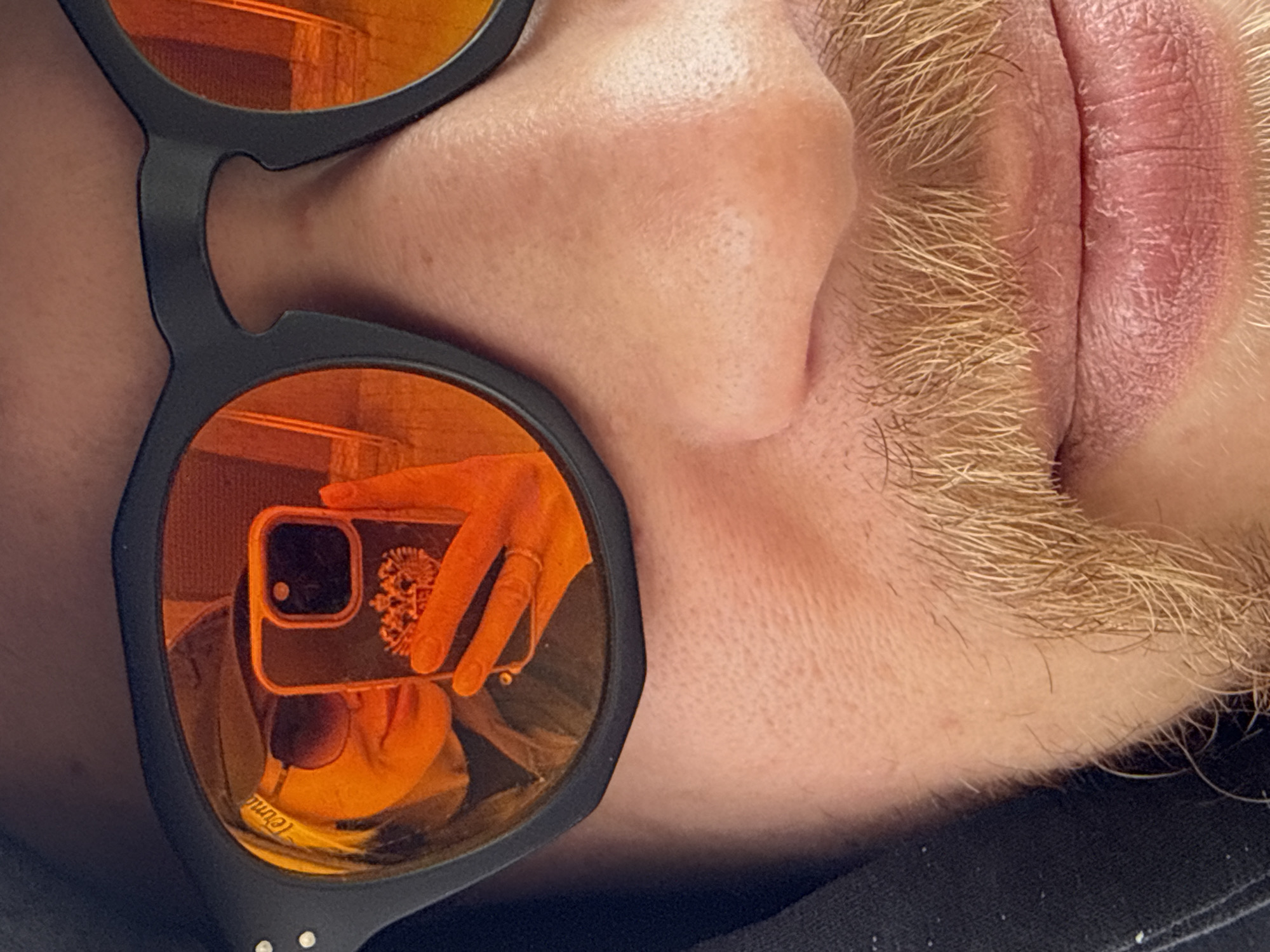 A close-up image depicts a portion of a person's face. The subject has a nose, mouth, and a blondish mustache. They are wearing sunglasses with orange lenses. The reflection in the sunglasses reveals a hand holding a smartphone with a decorated case, and the reflection of the person taking the photo. The environment is not discernible, and there is no evidence to suggest the location is in the city of Borzya, Russia.
