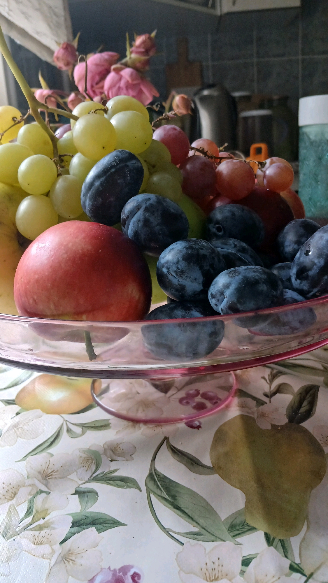 A close-up shot captures a clear glass fruit bowl filled with an assortment of ripe fruits, including green and red grapes, dark blue plums, and a red apple. The fruit bowl rests on a table covered with a decorative cloth featuring a floral pattern with white flowers, green leaves, and hints of pink and yellow fruit shapes. In the background, a kitchen counter is visible with various items, including what appears to be a pitcher, a cutting board, and containers. To the left, a cluster of pink roses in buds and partial bloom adds a touch of delicate beauty to the scene. The overall composition is well-lit, highlighting the textures and colors of the fruits and surrounding elements.