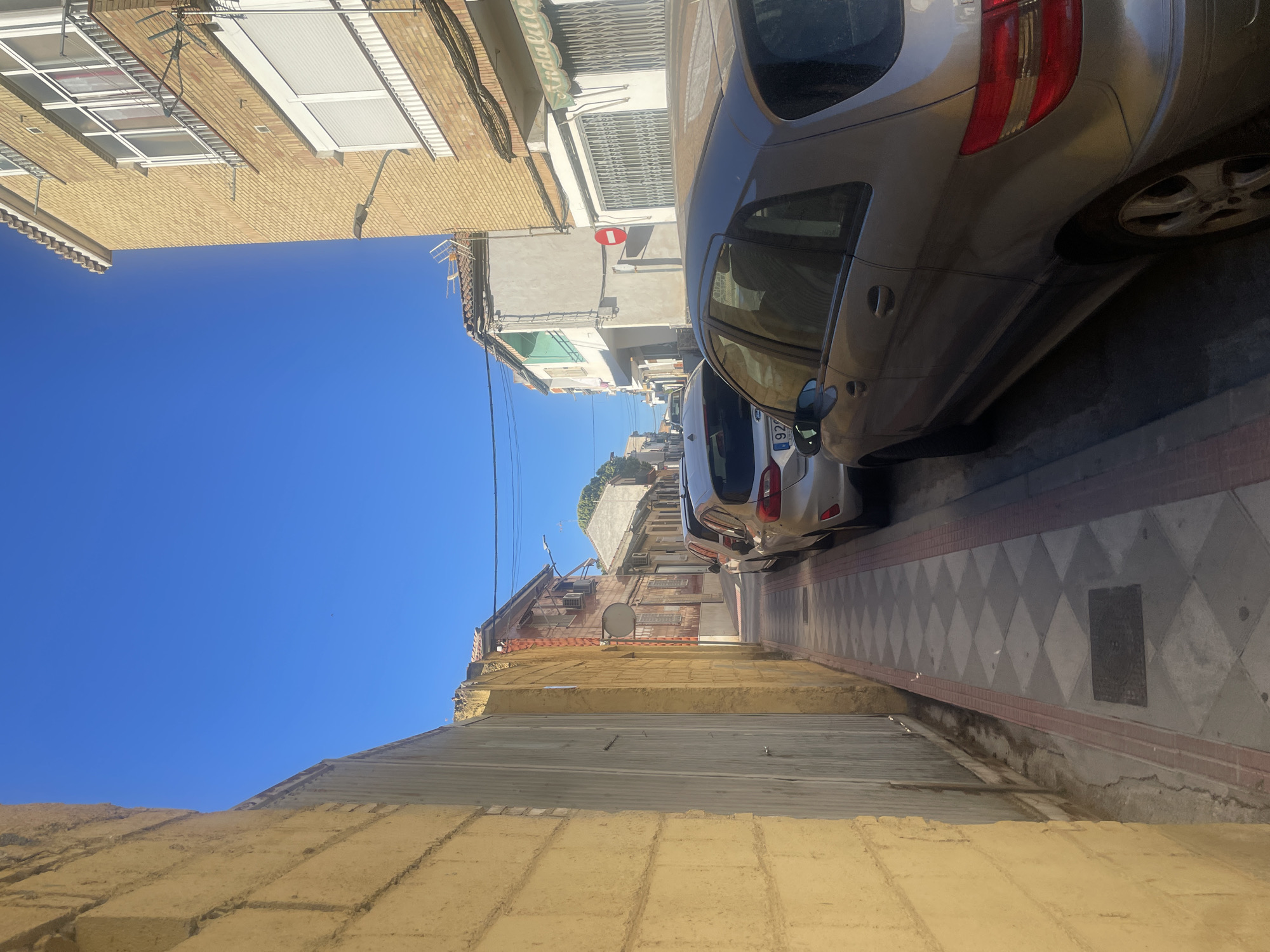 The image depicts a narrow street in Atarfe, Spain, lined with buildings and parked cars. On the left, a textured yellow brick wall forms the boundary of the scene, adjacent to a corrugated metal gate. The sidewalk is paved with a diamond pattern of light and dark grey tiles, bordered by red brick. Several cars are parked along the right side of the street, including a silver hatchback and a silver minivan. The buildings facing the street are a mix of architectural styles, with some featuring exposed brick and others painted in lighter colors. Balconies and windows are visible on the upper floors. The sky is a clear, bright blue, indicating a sunny day. The overall scene conveys a sense of an ordinary residential street in a Spanish town.