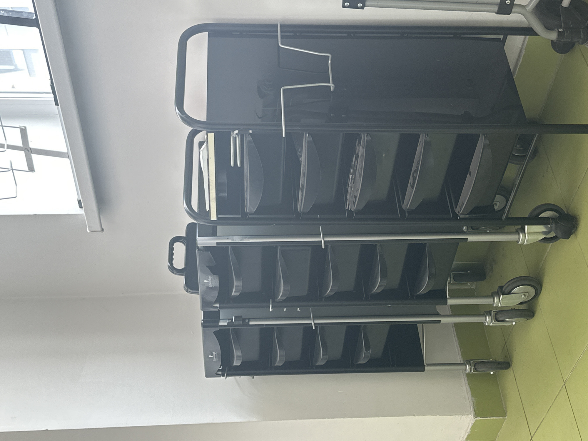 The image shows three black salon trolleys with multiple drawers arranged side-by-side in what appears to be a salon or similar commercial establishment. The trolleys have silver metal legs and wheels. The background consists of a white wall and a window with horizontal bars. The floor is tiled in a light green color. One of the trolleys has a rack attached to its side. The image is taken from a medium distance, showing the full height of the trolleys. There are no people visible in the image. The lighting suggests an indoor environment. The scene is clean and organized. The location is not specified as being in Moscow, Russia.