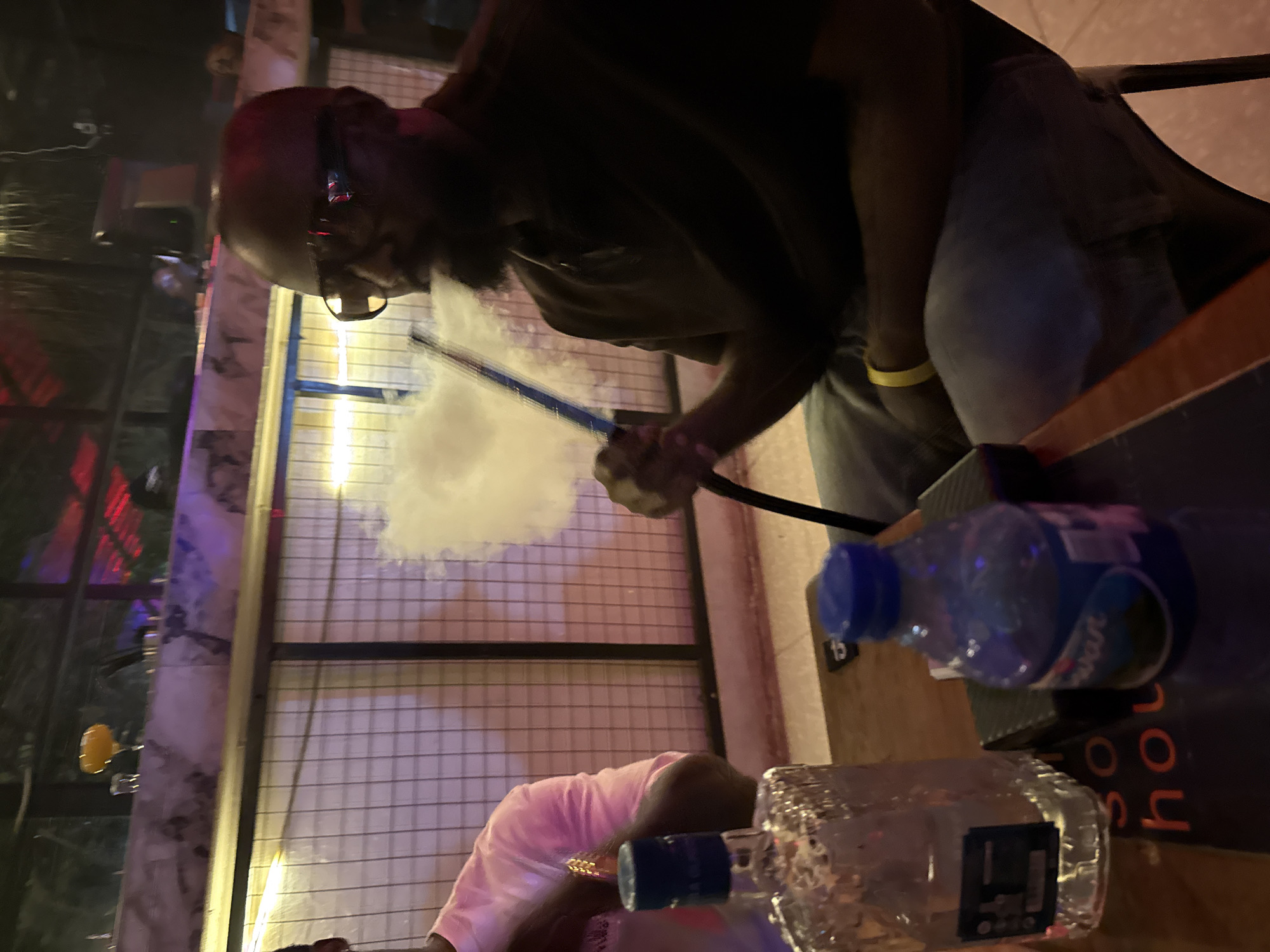 The image depicts an indoor or covered outdoor scene, possibly a lounge or restaurant, with a man in the foreground exhaling smoke from a hookah. He is wearing a dark shirt, jeans, and sunglasses, and has a beard. A bottle of clear liquid and a plastic bottle with a blue cap are on a table in front of him. To his left, another person is partially visible, wearing a pink long-sleeved shirt. The background features a patterned wall and some overhead lighting, with hints of red and purple illumination. The overall ambiance suggests a social gathering at night. The reference to Abuja, Nigeria, is not visually evident in the image itself.