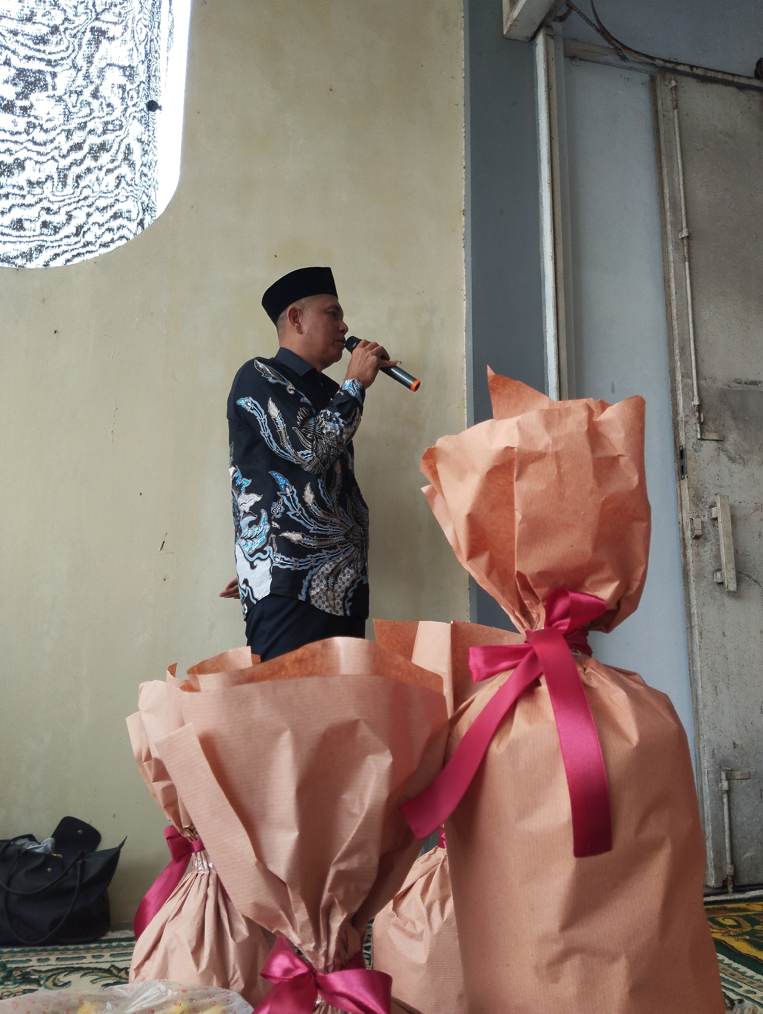 In Pangkah, Indonesia, a man wearing a black peci and a black patterned shirt stands facing left, speaking into a microphone. He is positioned in front of a cream-colored wall with a large, abstract, white and black patterned window on the left. In the foreground, two large gifts wrapped in light brown paper and tied with dark pink ribbons are prominently displayed. To the far left, a black bag is visible on the floor. The background includes a gray metal door and a portion of a patterned rug on the floor.