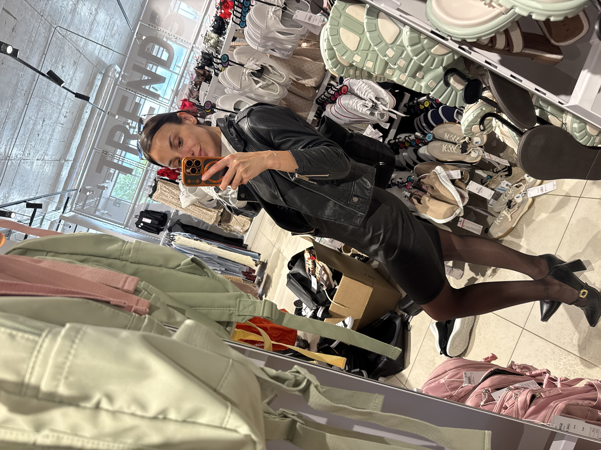 A woman wearing a black leather jacket and a black dress is taking a selfie in a clothing store. She is holding a phone with a brown case. Shelves of shoes are visible behind her, and clothing and bags are displayed on shelves in front of her. The store appears to be a "TREND" store, as seen in a mirrored sign on the wall. The setting is likely a shopping mall or large department store in Pleven, Bulgaria.