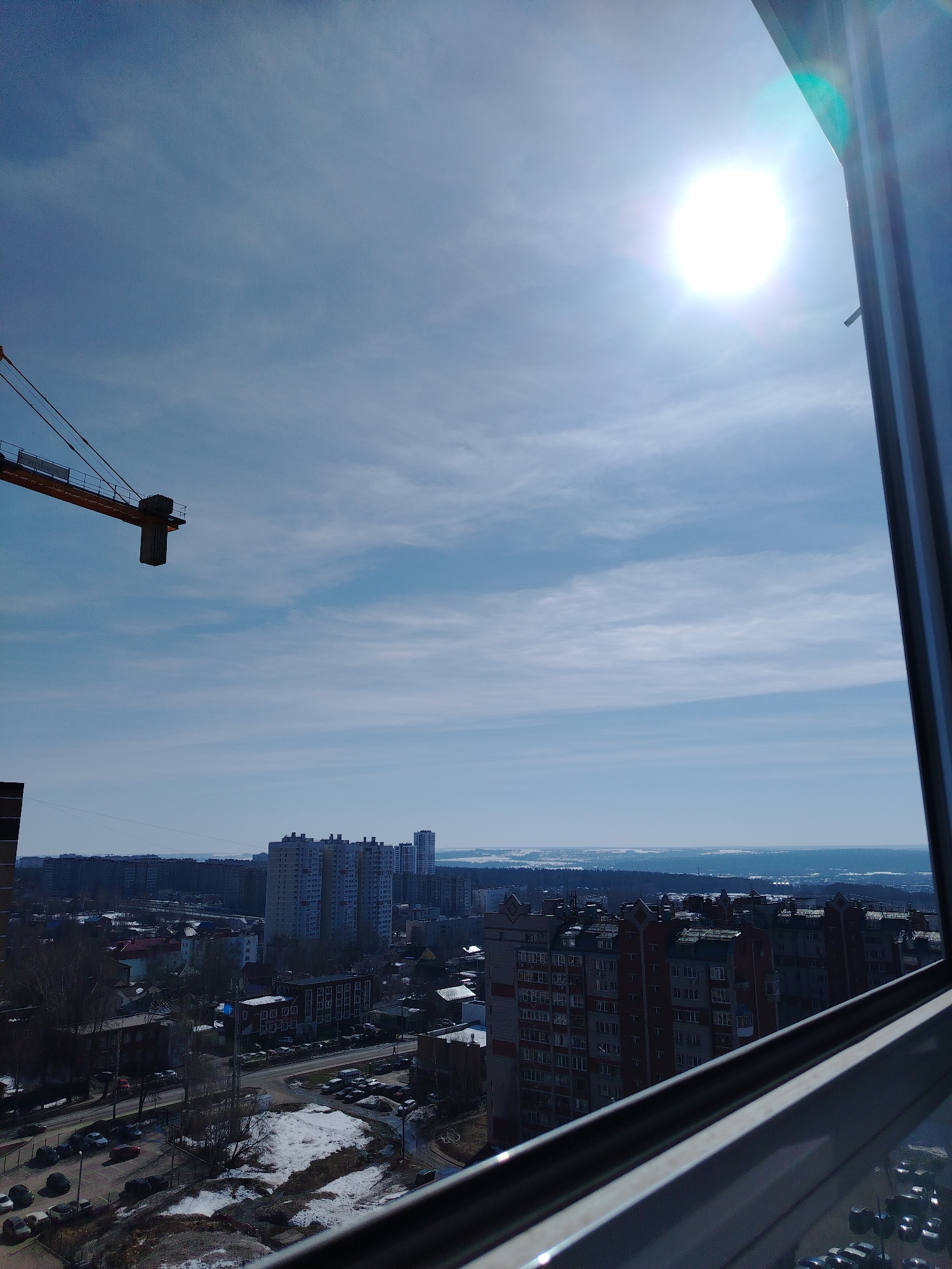 View from a window overlooking a city on a bright, sunny day. A construction crane is visible in the upper left. In the mid-ground are several high-rise apartment buildings. In the foreground, there is snow remaining on the ground. The location is most likely Izhevsk, Russia.