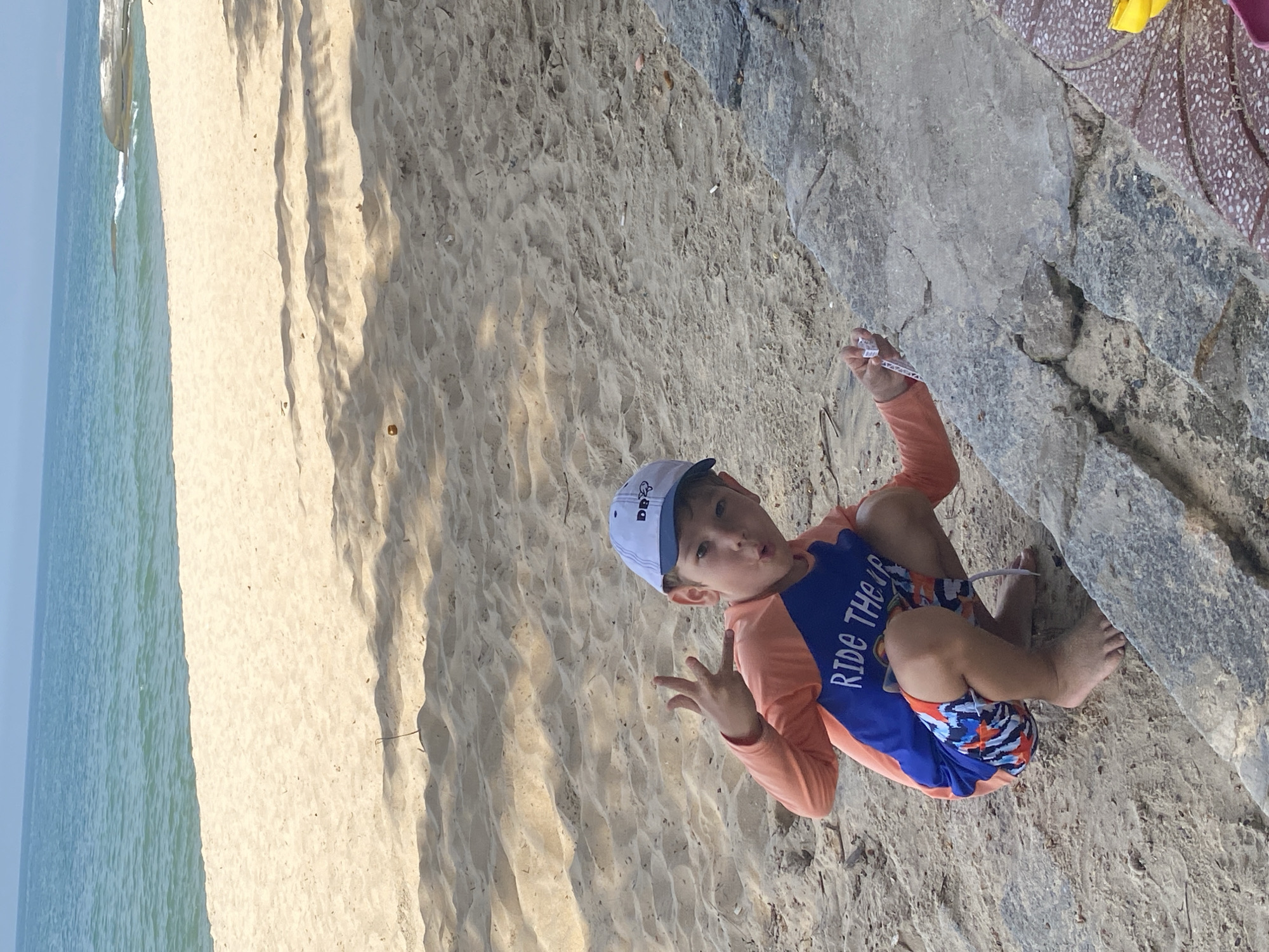A young boy wearing a blue and orange rash guard and swim trunks, and a blue and white baseball cap, is crouching on a sandy beach in Phu Thuy Ward, Vietnam. The boy is making a hand gesture with his fingers. He is looking at the camera and appears to be playing with a small white object in his hand. The beach has sand and rocks, and the ocean is visible in the background. The image is taken during the daytime, and there are shadows cast on the sand, indicating a sunny day.