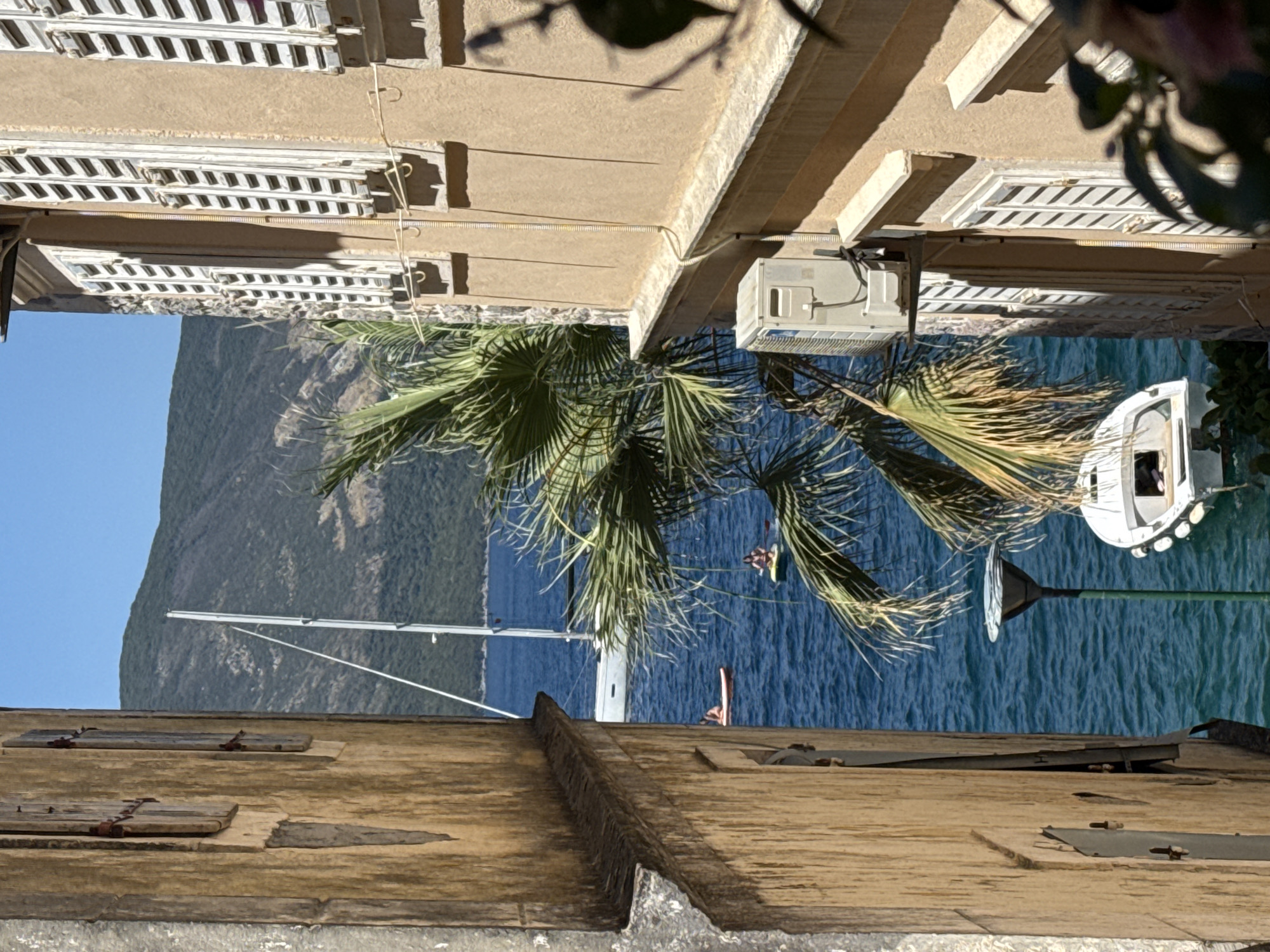 An outdoor, eye-level, medium shot captures a narrow alleyway opening to a bright, blue body of water. On the left, a rough, weathered, tan-colored building features wooden shutters with visible rust. On the right, a tan building with white shutters is visible, with an outdoor air conditioning unit mounted on its exterior wall. A palm tree, with green fronds, partially obscures the view of the water. In the background, a large sailboat with a white mast is anchored, and two people are visible on paddleboards in the water. A small white boat with a green light pole is docked near the shore. A verdant, tree-covered mountain range forms the distant backdrop under a clear blue sky.