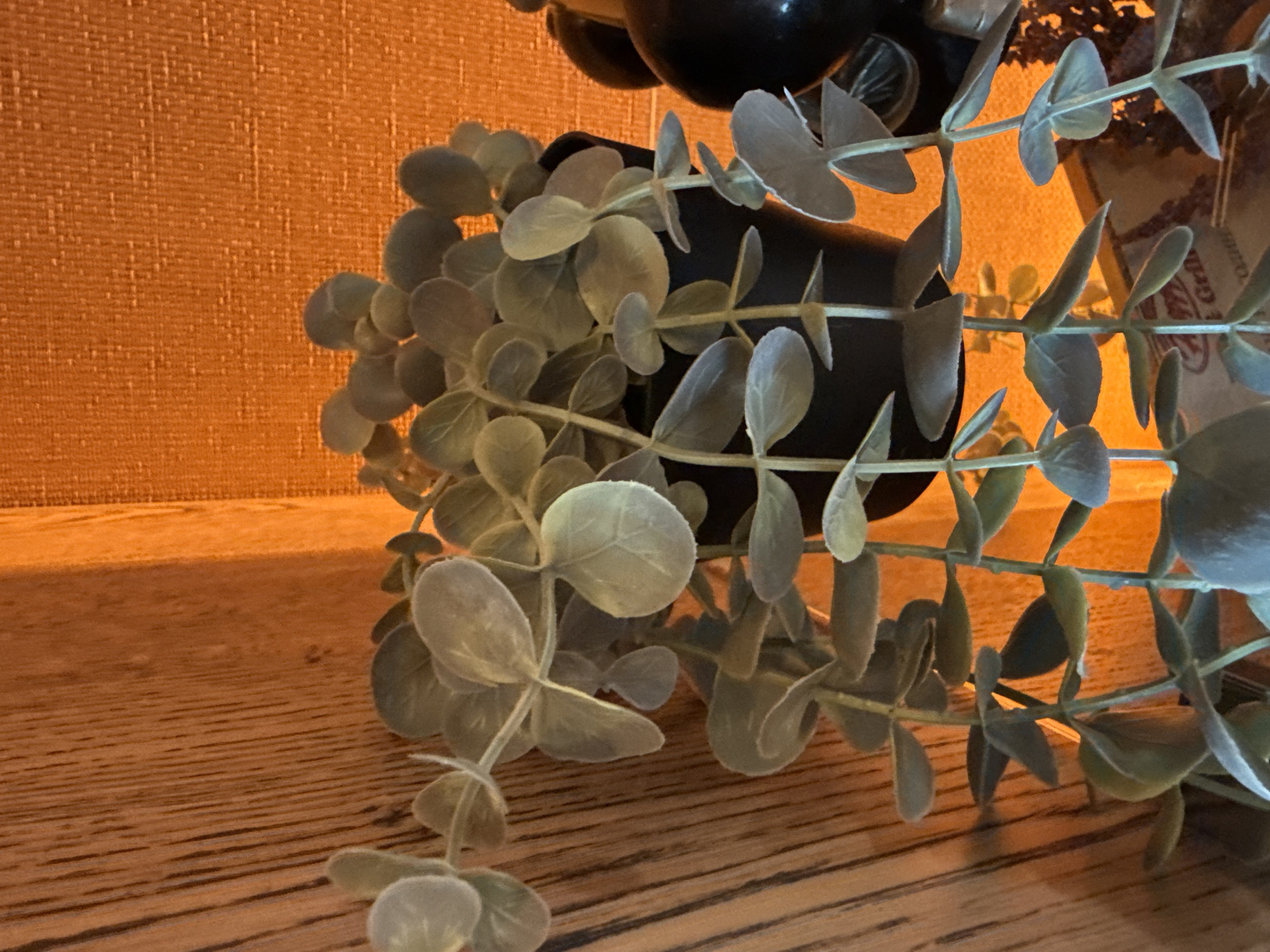 The image displays an indoor still life, focusing on an artificial plant and its immediate surroundings. The main subject is a faux plant, likely eucalyptus, featuring numerous small, rounded, grey-green leaves with a soft, slightly flocked texture. The plant's stems cascade from a dark, cylindrical pot, partially visible towards the top-center, and extend throughout the frame, particularly dense on the right side.
The plant rests on a warm-toned wooden surface, visible in the foreground and along the bottom-left, characterized by distinct horizontal grain patterns. Behind the plant, a wall or panel with a textured, possibly woven, surface glows with a vibrant orange hue, suggesting warm artificial lighting.
No activity or event is taking place; the scene is static. The lighting creates a cozy, intimate atmosphere. In the upper right background, partially obscured by the plant, there is faint, blurry text on a lighter surface. Visible elements of this text appear to include "Grill" and possibly "Radina" or "Radino", hinting at a location or establishment within Radinovo, Bulgaria. The overall impression is one of interior decor in a home or commercial setting.