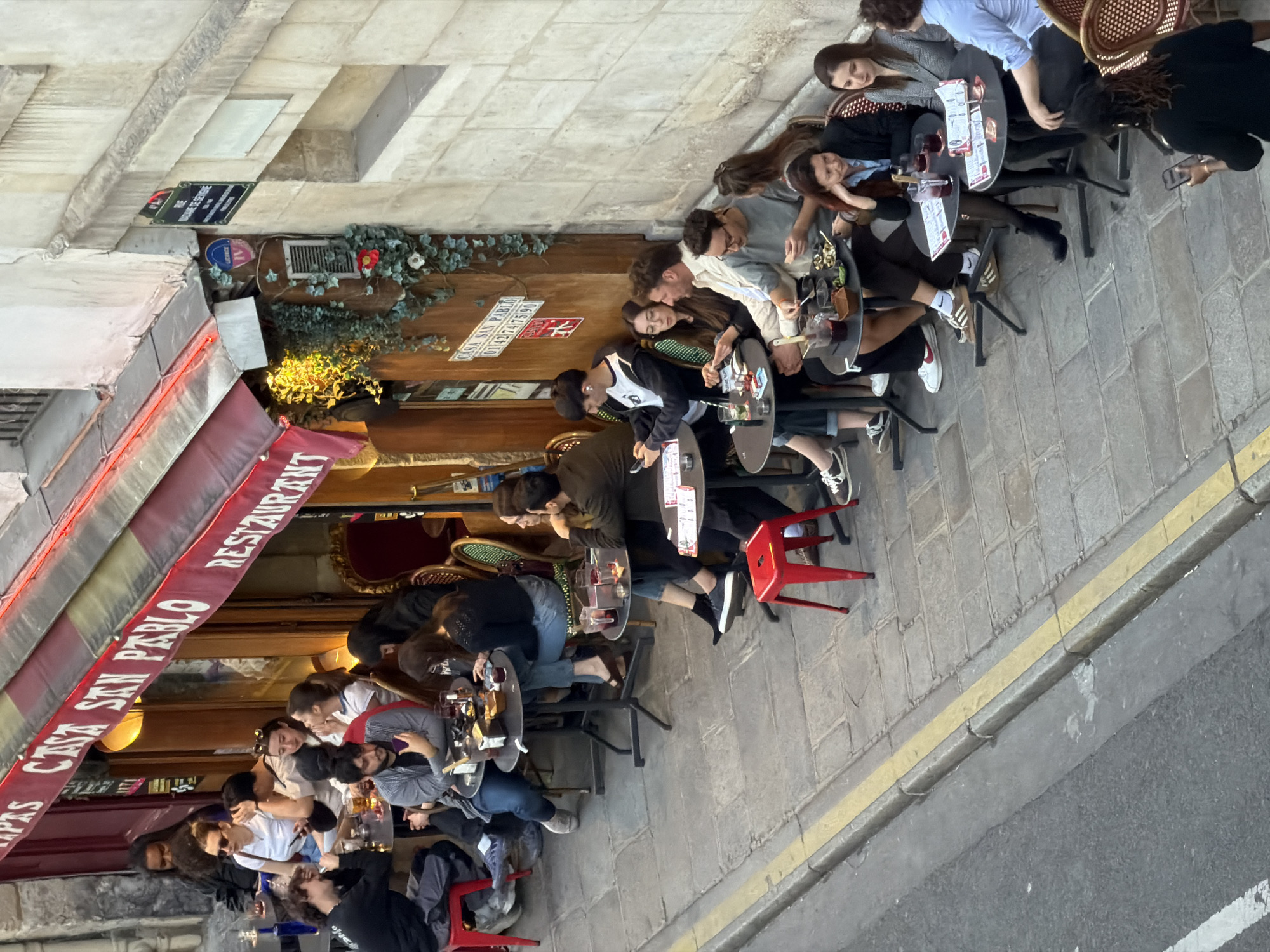The image presents an overhead view of a lively outdoor restaurant or tapas bar, "CASA SAN PABLO," situated on a narrow cobblestone street, likely in Paris, France. The street is identified by a visible black and white sign reading "RUE SAINTE-CROIX," with an additional sign showing the Roman numeral "IV," indicating an arrondissement.
The scene captures numerous young adults, casually dressed, socializing at closely-packed tables that line the sidewalk. They are engaged in conversations, using mobile phones, and enjoying drinks and small plates of food, suggesting a relaxed late afternoon or early evening gathering. The lighting is soft, with some artificial lights from the restaurant adding to the ambiance.
The restaurant features a distinctive red awning with "TAPAS CASA SAN PABLO RESTAURANT" written in white lettering. The building's facade is constructed from light-colored stone, adorned with ivy and other green foliage. A wooden panel near the entrance reiterates "CASA SAN PABLO" and displays a phone number, "0142747590." Seating comprises a mix of red plastic/metal chairs and traditional woven bistro chairs.
The street itself has a clear yellow line marking the curb along the cobblestone pavement, separating it from the darker asphalt road. People appear content and absorbed in their social interactions, creating a vibrant urban atmosphere.