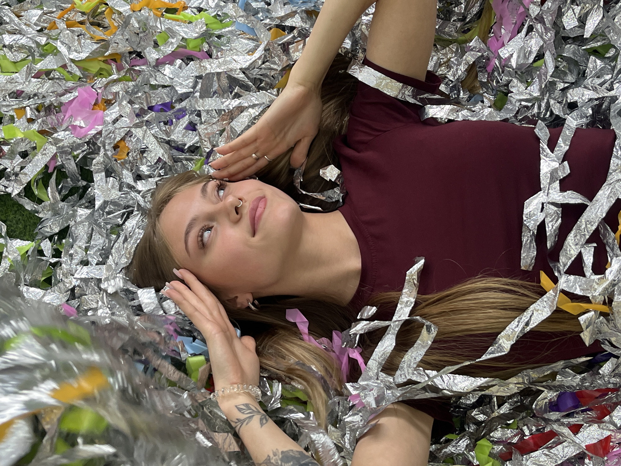 A young woman with light brown hair is lying on her back amidst a large quantity of metallic silver streamers or confetti. Her head is tilted slightly upwards, and she is looking away from the viewer with a calm, soft expression, a hint of a smile on her lips. Her hands are positioned near her head and face; one hand rests beside her head with two silver rings visible on her fingers, and the other is bent with the forearm visible, revealing a tattoo with a leafy design. She is wearing a maroon short-sleeved top.
The silver streamers are intricately wrinkled, reflecting light and creating a shimmering effect. Interspersed among the silver are various colorful pieces of paper or plastic, including vibrant pink, lime green, yellow, purple, blue, and red elements, suggesting a celebratory atmosphere. The setting appears to be indoors, possibly a party venue or a photo booth backdrop, given the abundance of confetti. There are no direct visual cues to pinpoint the exact location, but the context provided places it in Chaikovsky, Russia. The time of day and weather are indeterminate due to the indoor setting. No visible text is present in the image.