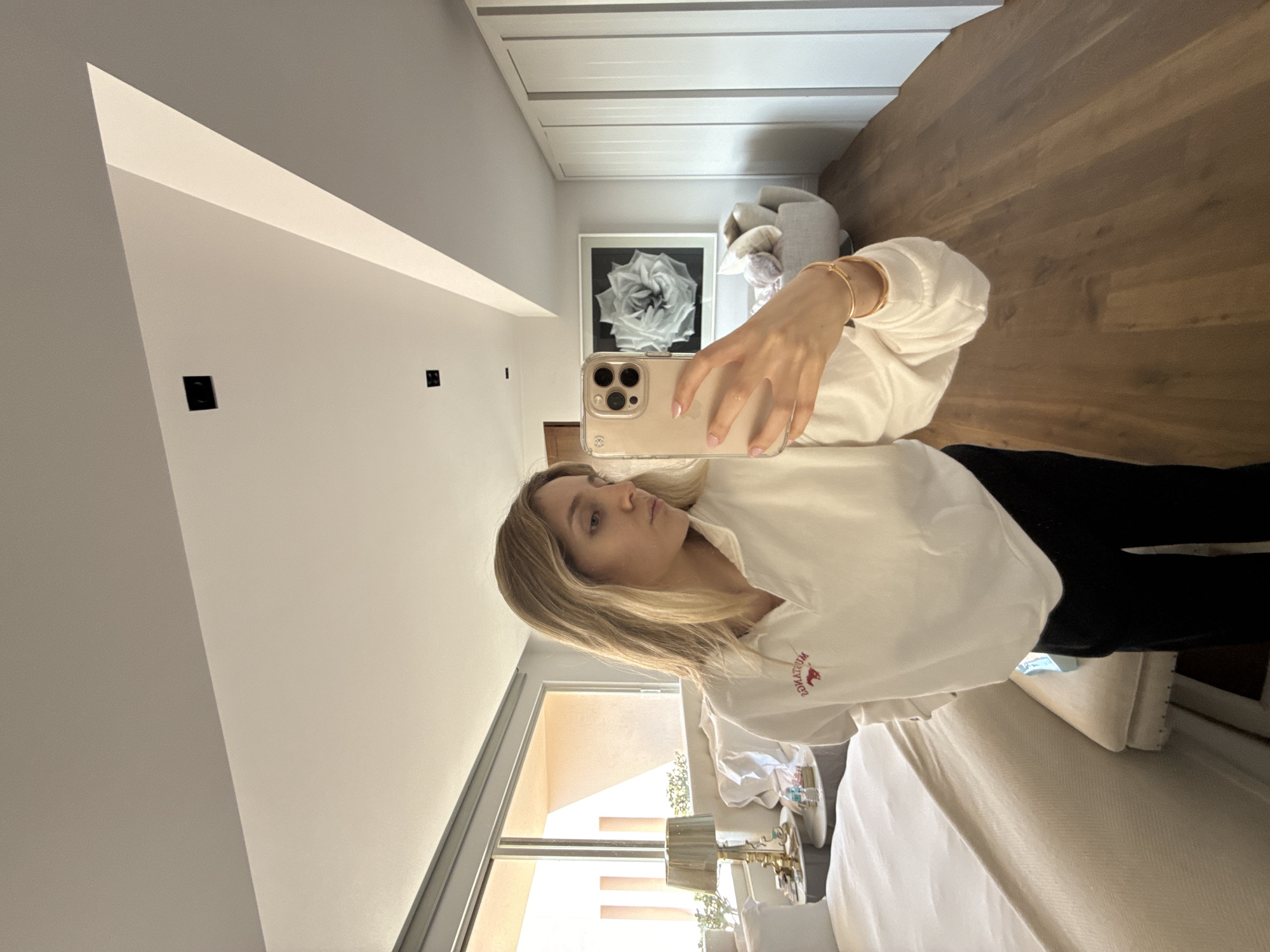 A woman is captured in a mirror selfie within a modern, well-lit room. She has blonde hair and is wearing a white long-sleeved top featuring a red "MOUNTAINUM" logo with a fox emblem, paired with dark pants. Her left hand holds a light-colored iPhone, encased in a clear cover, revealing light pink nail polish and a delicate gold bracelet on her wrist. Her expression is neutral as she looks towards her phone.
The interior showcases a contemporary aesthetic with predominantly white walls and a large reflective surface where the selfie is taken. The reflection reveals light-toned wooden flooring or ceiling and sleek white built-in cabinetry or doors. A prominent framed artwork depicting a large, detailed white rose in black and white hangs on one wall, with a light-colored armchair or sofa positioned nearby. Two black square electrical outlets are visible on the wall.
Through a large window or sliding glass door, a bright and sunny outdoor scene can be seen, characterized by light-colored building structures and some lush greenery. Near the window, a bedside table holds a lamp and other small items. The bright natural light indicates it is daytime. The scene suggests a clean, modern, and comfortable indoor space. The location context for this setting is Mexico City, Mexico.