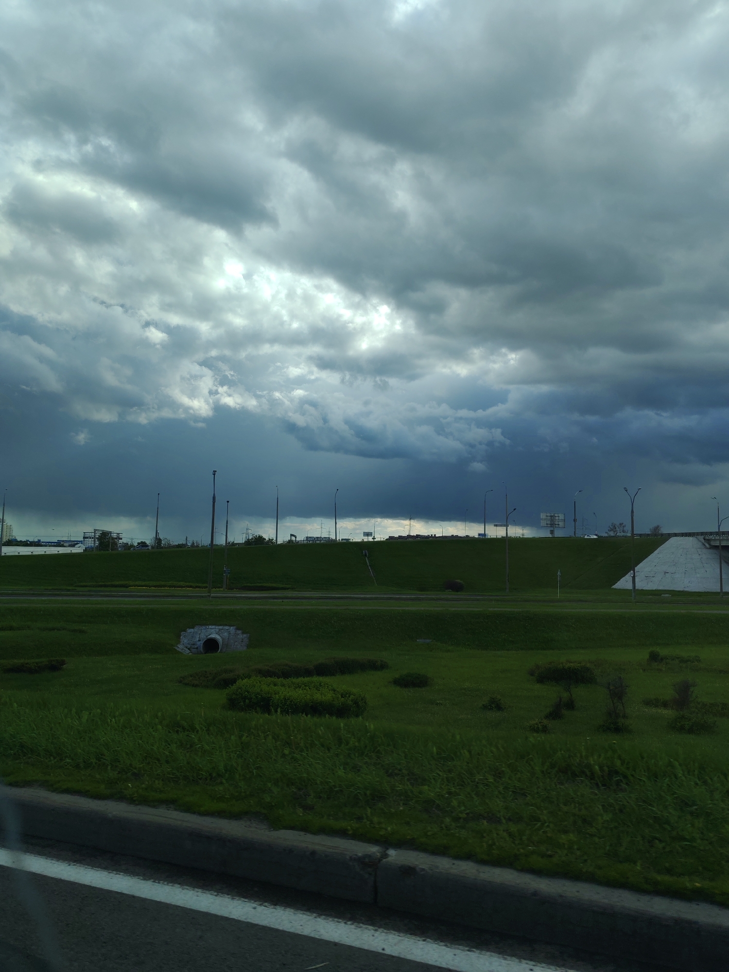The image presents a landscape under a dramatic, overcast sky. The sky is filled with dark, heavy clouds, suggesting an imminent storm. The ground is covered in lush green grass, forming a field or park-like area. A concrete culvert is visible in the middle ground. The background features a raised area, possibly a hill or embankment, with streetlights lining the top. On the embankment is a small white concrete structure on the far right. The presence of streetlights and what appears to be a road suggests an urban or suburban setting. A road is visible in the foreground. The location is in the city of Unknown, Unknown.