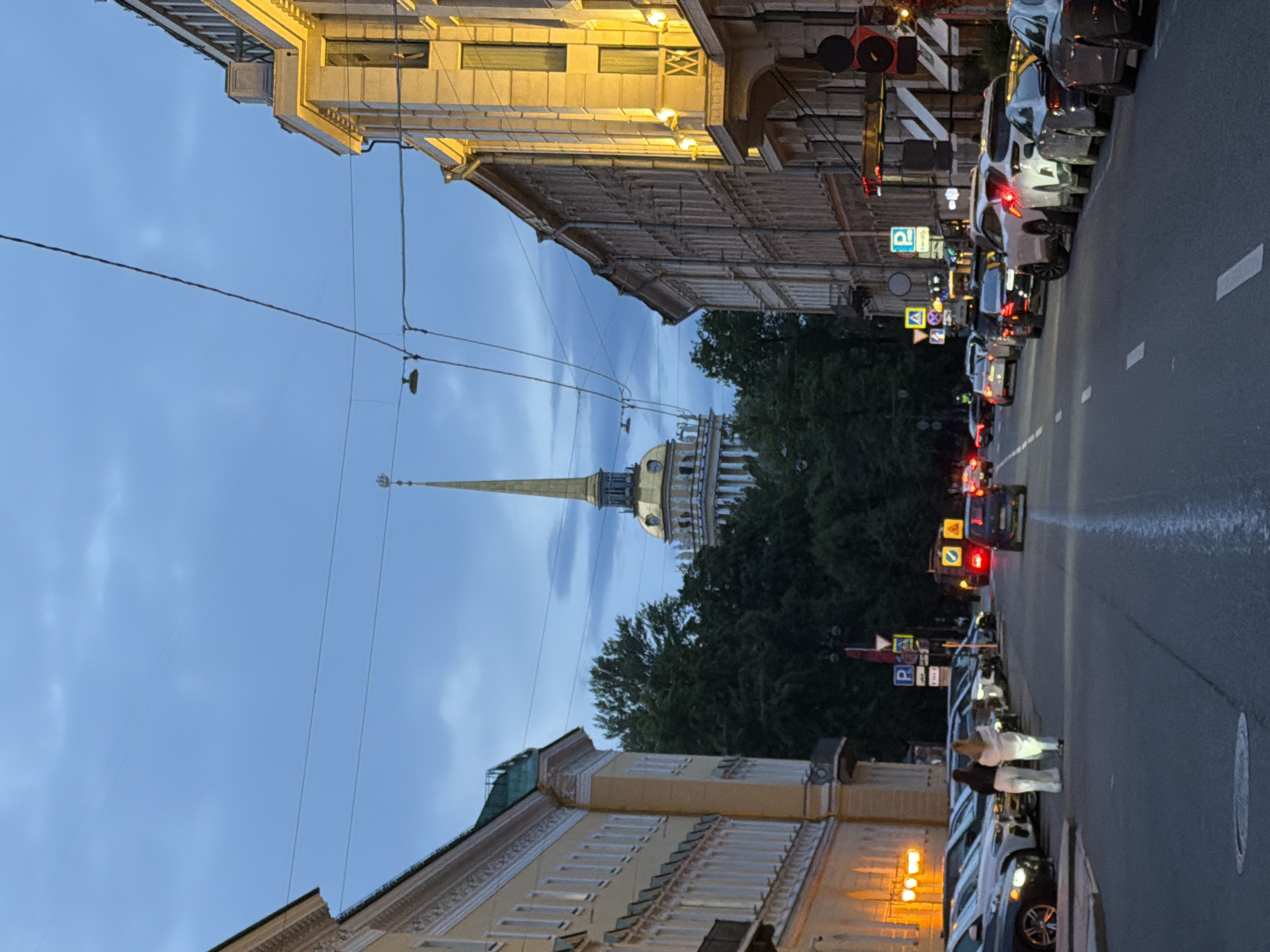 The image depicts a street scene in Saint Petersburg, Russia, at dusk or early evening. The perspective is from street level, looking up towards a tall spire of a building, likely the Admiralty Spire, topped with a golden dome. The spire rises above a row of trees and adjacent buildings. The sky is a muted blue with hints of clouds, suggesting twilight.
On the right side of the frame, a street is lined with parked cars. Some of the cars have their brake lights illuminated, indicating that traffic may be moving or stopped. Traffic lights and street signs are visible, including a "P" sign for parking. The buildings flanking the street are multi-story, with architectural details suggestive of European cityscapes. One building on the left has ornate detailing and illuminated windows.
A few people are visible near the parked cars, walking on the sidewalk. They appear to be casually dressed. The overall mood of the image is calm and somewhat serene, capturing the transition from day to night in an urban environment. The lighting suggests it is not yet fully dark, with some streetlights and building lights beginning to cast a warm glow. The presence of overhead wires indicates the infrastructure of the city.