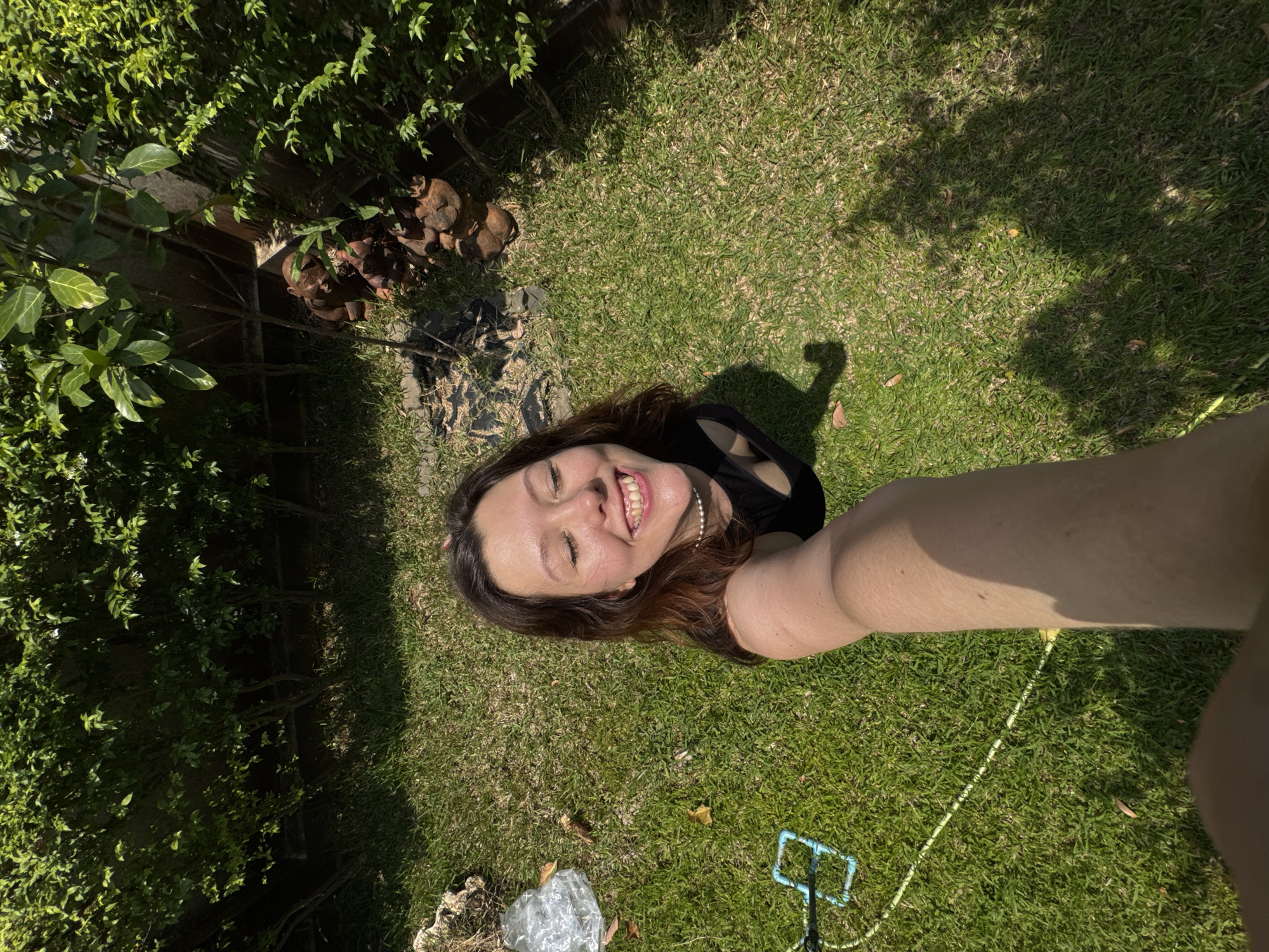 The image shows a woman taking a selfie in a grassy area outdoors in บ้านกาญจน์กนก 23, Thailand. The woman is smiling with her eyes closed, facing the camera. She has brown hair and is wearing a black top and pearl earrings. Her arm is extended towards the camera. The grassy area is partially shaded by trees, and some objects are lying on the grass: a plastic bag and a blue plastic object connected to a yellow string. A bush and a collection of rocks are visible in the background.