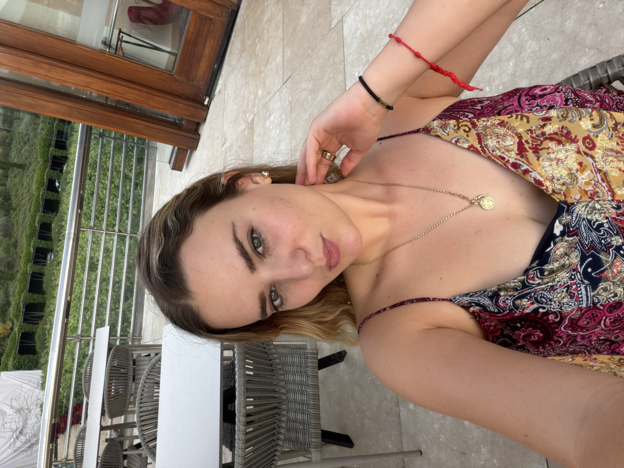A young woman with fair skin and wavy blonde hair poses in what appears to be a selfie outdoors. She is wearing a multi-colored, patterned dress with thin straps. She has a gold chain necklace and multiple bracelets on her wrist. There are tables and chairs visible to the left, suggesting a patio or balcony. Behind the railing is a green, plant-covered structure, and there is a building visible in the background. The location is potentially in Elciego, Spain, based on the architectural style of the building.
