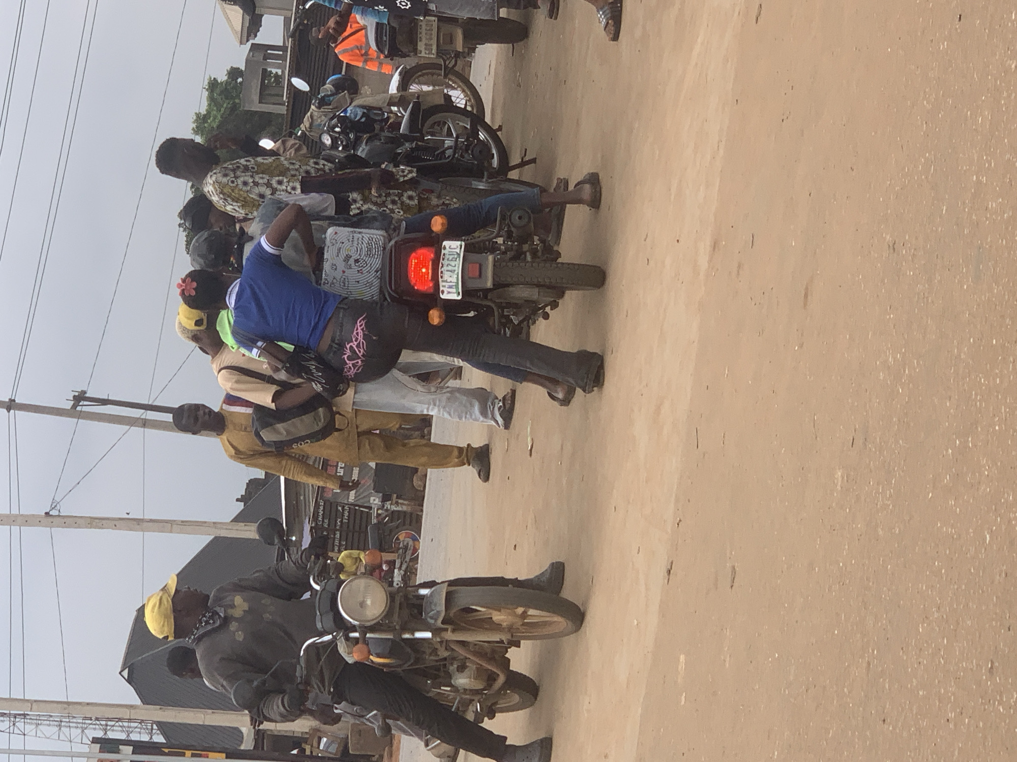 In Shagamu, Nigeria, a group of people are gathered around motorcycles on a dusty, unpaved road. In the foreground, a man wearing a yellow cap and a grey long-sleeved shirt is seated on a black motorcycle, facing left and looking forward. To his right, another man in a yellow traditional outfit stands with a backpack slung over his shoulder. Behind them, a woman in a blue shirt and patterned leggings is seated on the back of a motorcycle, with her arms around the person in front of her. Other individuals are standing or seated on motorcycles in the background. The environment appears to be an urban setting with power lines visible overhead and simple structures in the distance. The ground is covered in reddish-brown dirt, indicating a dry, possibly arid climate. The overall scene depicts a moment of transportation and social interaction amidst the everyday life of Shagamu.