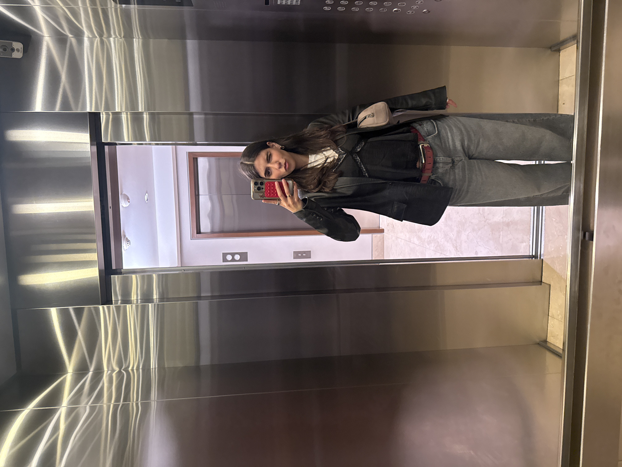 The image shows a young woman taking a selfie in an elevator. The elevator appears to be in a modern building with mirrored surfaces. The woman is wearing jeans, a black top, a black jacket, and a red belt, and she has a small beige bag. She is holding a red phone in her left hand. The background of the image through the elevator doors reveals a hallway with light-colored tile flooring and white walls. The scene may be in Mexico City, Mexico.