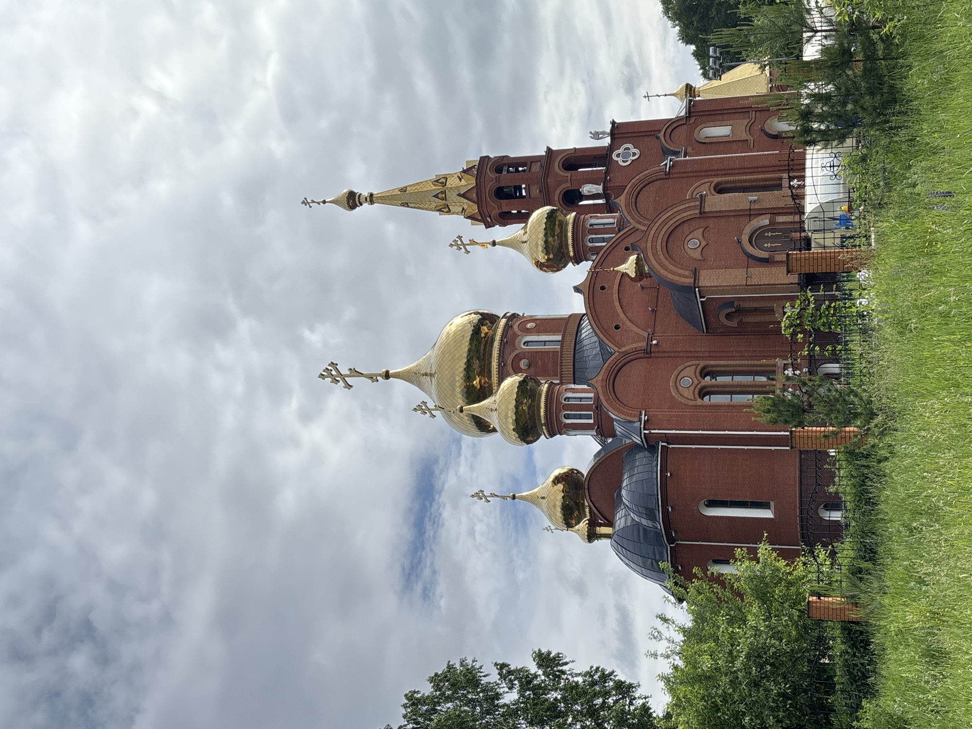The image captures a large, ornate Eastern Orthodox church, located in Chaikovsky, Russia. The church is constructed from red brick and features multiple golden, onion-shaped domes of varying sizes, each topped with a characteristic Orthodox cross. A tall bell tower with a golden spire and cross is prominent on the right side of the structure. Architectural details include arched windows and decorative elements on the brickwork.
The scene is set amidst green natural surroundings, with lush grass and various trees and shrubs in the foreground and around the church perimeter. A dark metal fence with decorative ironwork partially encloses the church grounds.
The sky is overcast with light gray clouds, indicating a cloudy daytime setting. No individuals or specific activities are depicted in the frame, emphasizing the architectural subject. There is no visible text within the image. The overall impression is one of architectural grandeur and peaceful solemnity.
