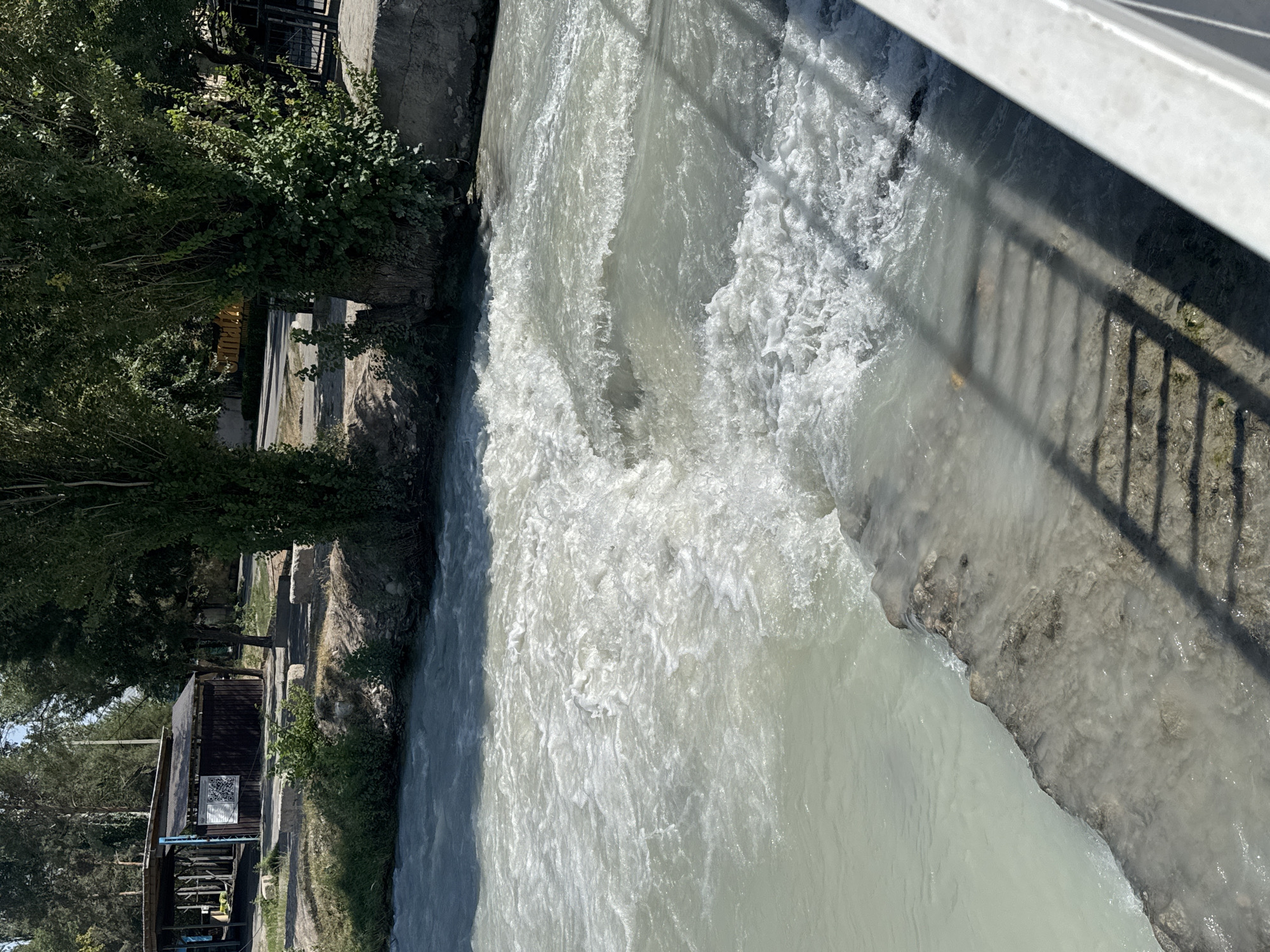 The image captures a view of the Ala-Archa River in Bishkek City, Kyrgyzstan, on a sunny day. The river flows rapidly over a low dam or weir, creating white water and rapids. On the left bank, there is a covered structure with a dark brown wooden building behind it, possibly a small cafe or rest area, and some vegetation. Trees with lush green foliage line the banks. A concrete embankment with a metal railing is visible in the foreground on the right, casting a shadow of the railing onto the water. The water itself appears milky or turbid, common for glacial rivers in mountainous regions. The overall scene depicts a natural environment within an urban setting, with the river being the dominant feature.