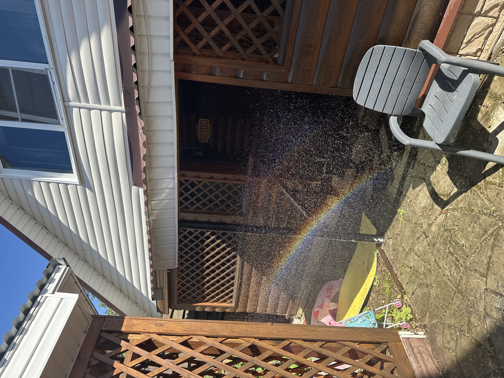 A rainbow is visible in the spray of water in front of a wooden building with white siding and a corrugated roof. A grey plastic chair is in the foreground, and a stone patio is underfoot. A child's toy stroller and a yellow mat are on the ground to the left of the building. The image was taken in the city of Nizhny Novgorod, Russia.
