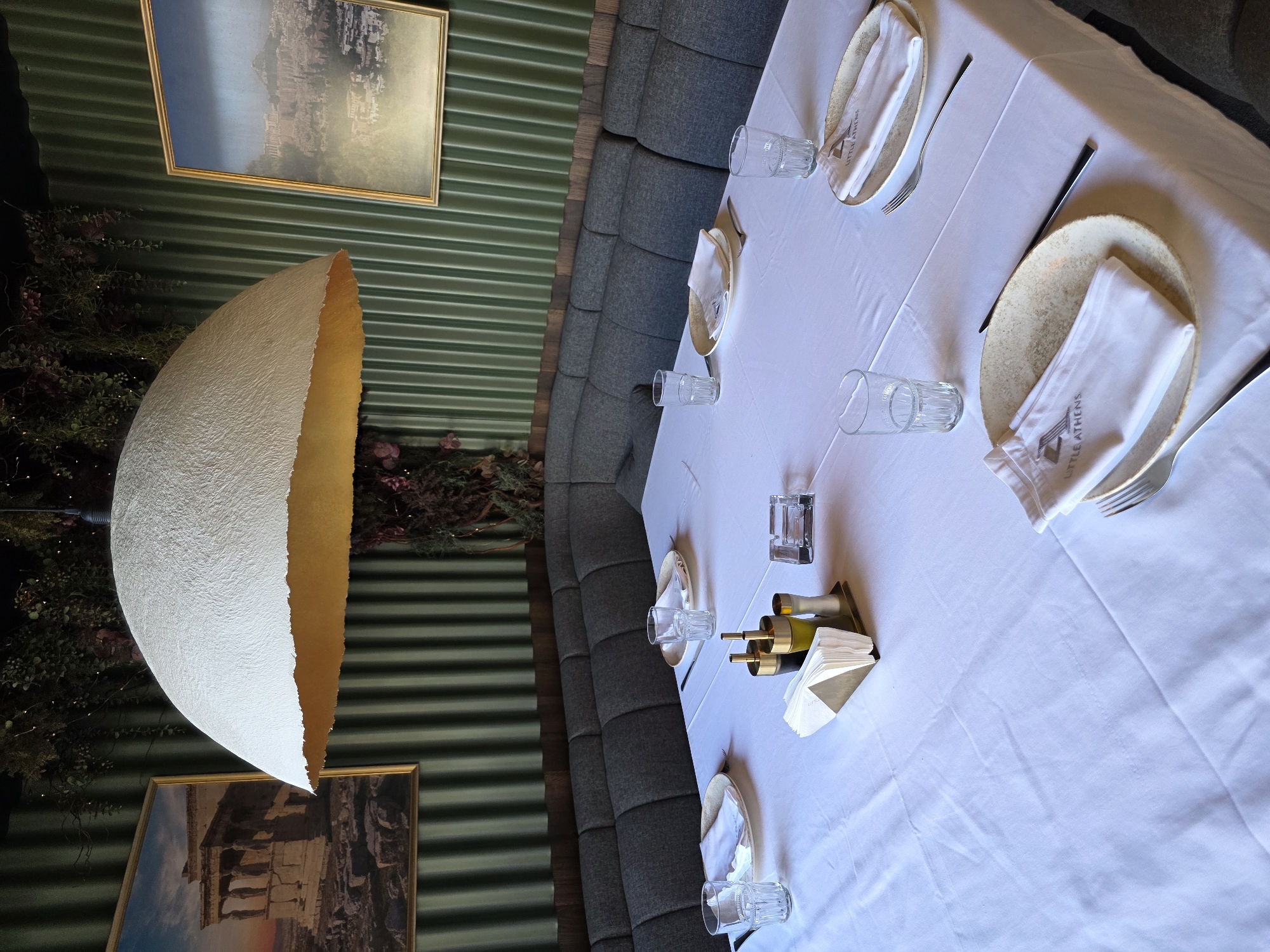 The image shows a dining table set for a meal in a restaurant. The table is covered with a white tablecloth and set with plates, cutlery, and glasses. There are also small bottles of what appear to be olive oil and vinegar. In the background, a curved banquette seat lines a wall covered in green corrugated material. Two framed pictures hang on the wall, one depicting a classical architectural landscape and another showing a similar scene. A large, egg-shaped light fixture hangs from the ceiling, casting a warm glow. The overall ambiance appears to be modern and elegant, with a focus on sophisticated decor. The lighting suggests it might be evening or an indoor setting. There is no visible activity or people in the image. The text "LITTLE ATHENS" is visible on the napkins.