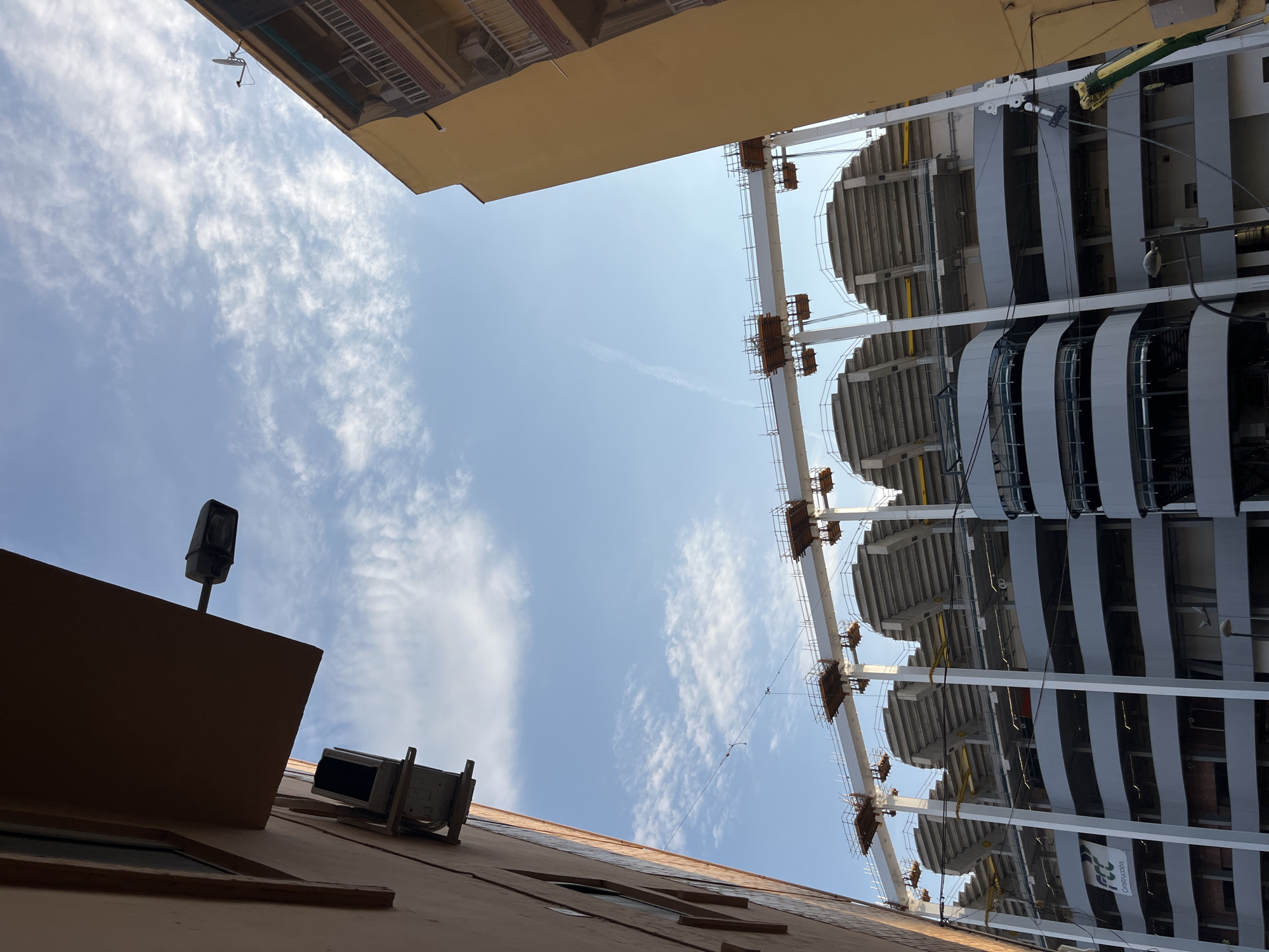 This media file shows an upward-looking perspective of a building under construction or renovation. The scene is dominated by the sky, filled with scattered white clouds against a clear blue backdrop, suggesting a bright, sunny day. The time of day appears to be late morning or early afternoon due to the lighting conditions.
On the left side of the frame, the edge of a building with a light brown facade is visible. It features a dark rectangular lamp attached to a pole extending from the roof, and an air conditioning unit mounted on the wall below. The building's structure is angled, creating a sense of looking up from a narrow street or alley.
The right side of the image is dominated by a large, multi-tiered structure, likely a stadium or a large public building, in various stages of construction. White support beams and horizontal beams form the framework, supporting rows of concrete seating. Gray curved panels are being installed on the exterior. Construction elements such as scaffolding and temporary supports are visible. A banner with the text "FCC Construcción" is attached to one of the beams on the lower right.
The overall impression is one of ongoing development and urban infrastructure. The contrasting elements of the older building facade and the modern construction suggest a dynamic urban environment. No people are clearly visible in the frame.
