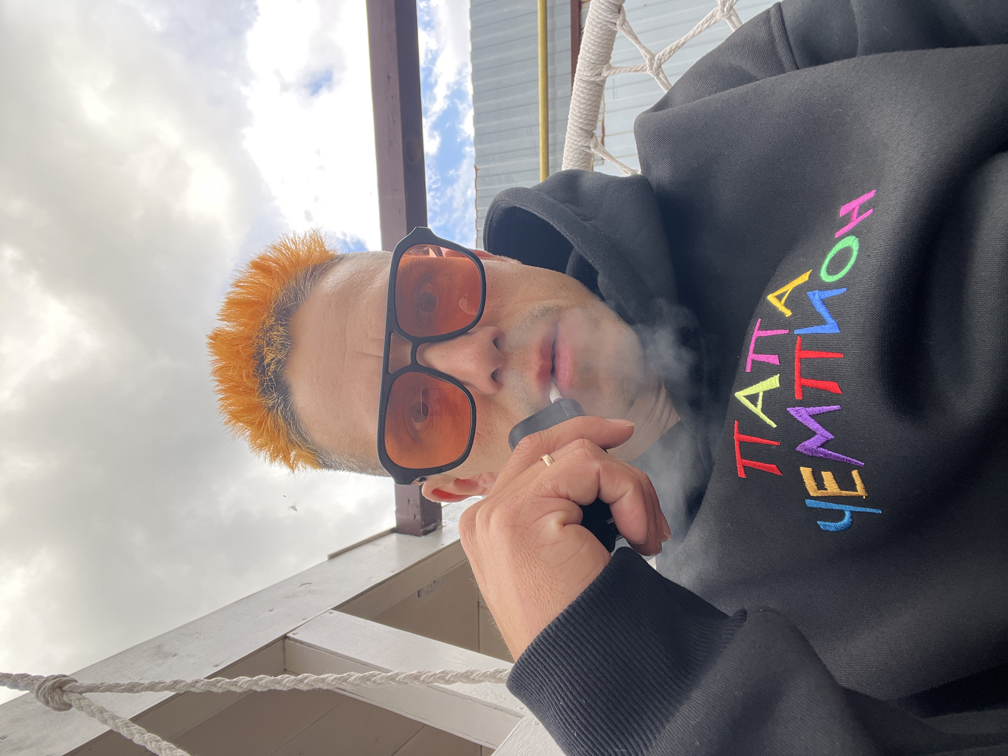 This media file is a close-up, low-angle selfie of a person with brightly dyed orange hair. They are wearing black sunglasses with orange lenses and a black hooded sweatshirt. The sweatshirt has colorful embroidered text on the chest, partially visible as "TA HEM". The person is holding a black e-cigarette to their mouth, exhaling a puff of smoke. They are also wearing a thin gold ring on their left ring finger.
The background suggests an outdoor setting, possibly a porch or balcony, with wooden structures and white rope visible. The sky is overcast with some patches of blue sky and clouds, indicating daytime. There are hints of industrial structures in the distance. The overall mood appears casual and relaxed. The visible text on the sweatshirt is likely a brand name or slogan.