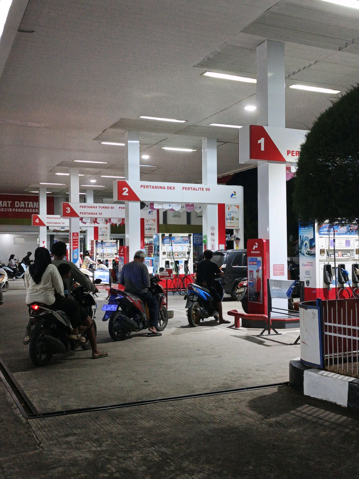 The image depicts a gas station in Pangandaran, Indonesia, at dusk or night. The gas station features multiple white pillars supporting a large roof, illuminated by fluorescent lights. Red and white signage indicates different fuel types, with prominent numbers 1, 2, 3, and 4 visible on some signs. Several fuel pumps are lined up, some of which are being used by customers.
In the foreground, three individuals are seated on motorcycles. The person on the left is wearing a black hijab and a cream-colored top, while the person behind them is in a grey long-sleeved shirt. In the center, a person in a blue shirt and dark pants is on a blue motorcycle. To the right, a person in a dark shirt and dark pants is seated on a red motorcycle. The ground is paved concrete, and shadows are cast by the structures and vehicles.
In the midground, more motorcycles and a dark-colored SUV are present. Some people are standing near the fuel pumps. A large banner with text in Indonesian is visible on the left side of the image. The background shows more fuel pumps and displays for various products. A dark green tree or shrub is visible on the right side of the image, partially obscuring the view. A white and red striped fence is also present in the right foreground. The overall impression is a busy, functional gas station environment in Indonesia.