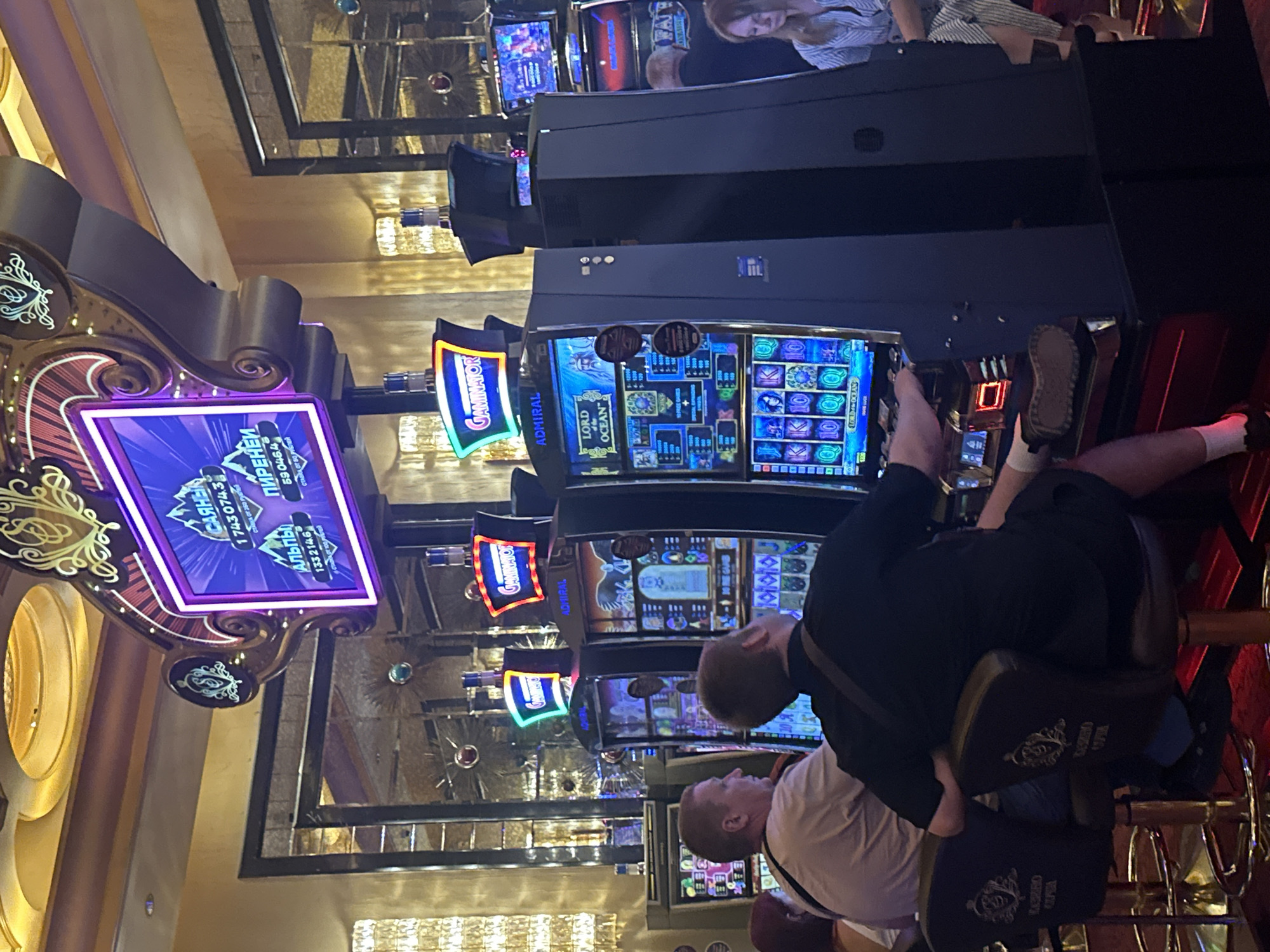 In the city of Estosadok, Russia, a dimly lit casino floor is captured in an image. The scene is dominated by rows of slot machines, their screens illuminated with colorful graphics and text in Cyrillic. One prominent machine displays "GAMINATOR" in glowing letters. In the foreground, two men are seated at bar stools in front of slot machines, with one man operating a machine, his hand resting on the control panel. Both men are casually dressed. A woman is visible in the background, also seated, her face partially obscured. The casino environment features ornate decorative elements, including mirrored walls and decorative lighting fixtures, contributing to a luxurious ambiance. The floor appears to be carpeted in red.