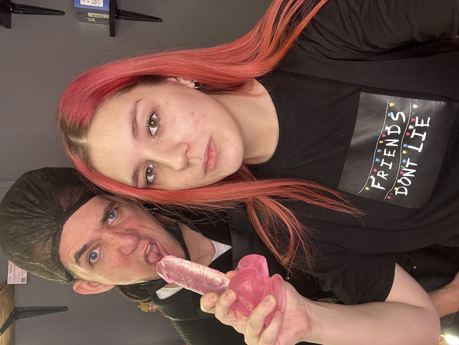 An indoor portrait photograph features two individuals, a female and a male, in what appears to be a commercial or private setting in Saratov, Russia, evidenced by Cyrillic text on a background sign.
The female, positioned on the right and facing the camera, has long, reddish-pink hair with visible dark roots. She wears a black long-sleeved t-shirt displaying the white graphic "FRIENDS DON'T LIE" adorned with colorful string lights. Her expression is neutral. Her right hand firmly grasps the flared base of a translucent pink phallic-shaped object, consistent with a dildo, holding it towards the center of the frame.
The male, situated behind and to the left of the female, wears a black disposable hairnet over dark hair, a black outer garment, and a white undershirt. His blue eyes are wide, and his facial expression is exaggerated, with his mouth agape and tongue extended, appearing to interact with the tip of the pink object.
The background consists of dark walls. A white rectangular sign with Cyrillic text, including "Придумал" (Invented), and a QR code is partially visible above the male's head. Black wall-mounted shelves containing small items are present in the upper right background. A golden decorative bust is also partially visible behind the male's left shoulder. The scene is illuminated by artificial lighting.