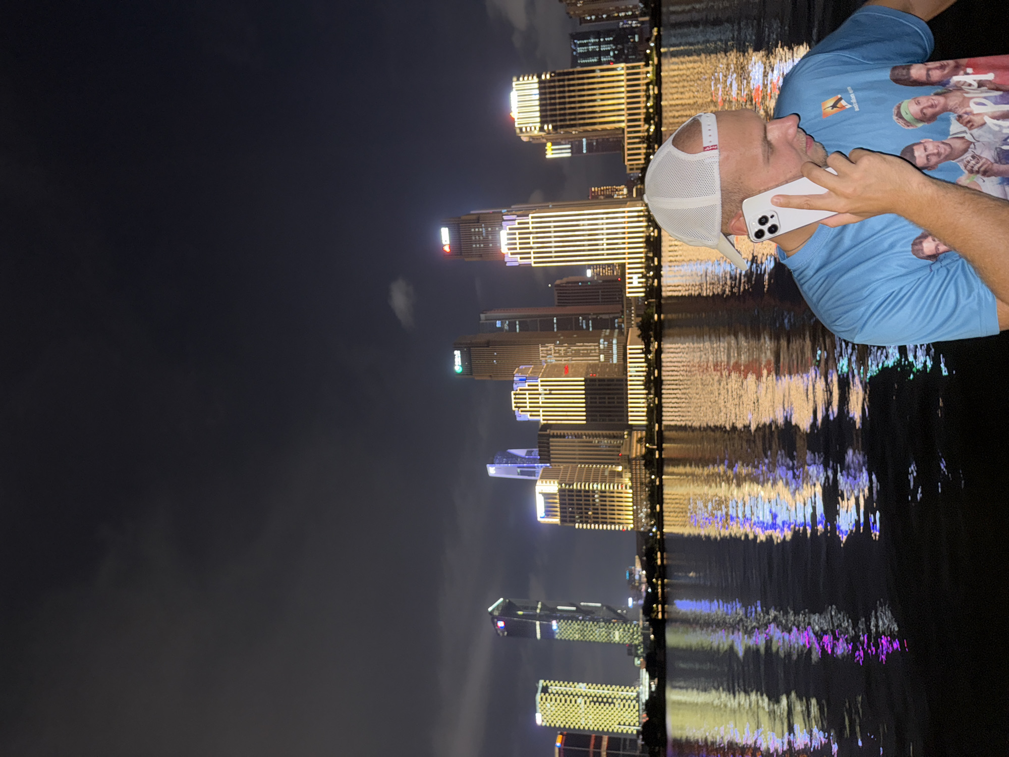 The image depicts a nighttime urban landscape with a male individual in the foreground. In the lower right, a Caucasian male wears a light blue t-shirt featuring an orange logo and a graphic of four individuals, along with a white mesh baseball cap worn backward. He is seen holding a white smartphone to his right ear, looking downwards and to his right, engaged in a call. Behind him, a dark body of water reflects the illuminated buildings of a city skyline. The reflections appear as vertical streaks of gold, yellow, purple, and blue light. The background features a dense collection of modern high-rise buildings along the water's edge, primarily illuminated with bright yellow and gold lights, some with blue and green accents. The sky above is dark with visible cloud formations. The scene is consistent with a waterfront view in a major city, specifically identified as Haizhu District, China, at night.