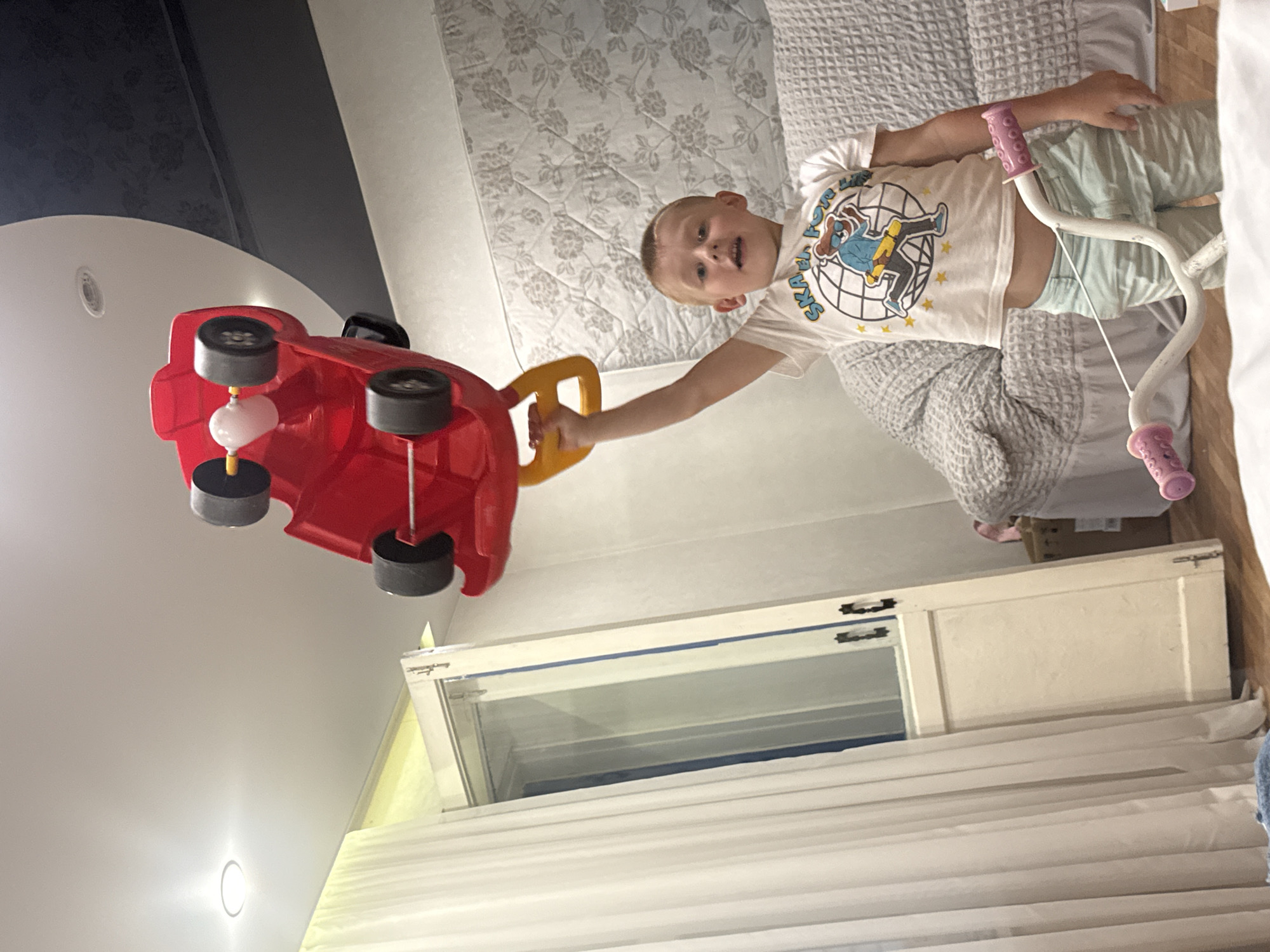 A young boy is standing in a room, holding a red toy car over his head. The boy is wearing a white t-shirt with a graphic print and light blue shorts. He is smiling and looking at the camera. The toy car appears to be upside down, with its wheels pointing towards the ceiling. The room has a light-colored ceiling with recessed lighting, and a window with white curtains on the left side of the frame. There is a bed with a patterned cover in the background. The floor is wooden. The scene is likely indoors, in a residential setting, in the city of Valday, Russia.