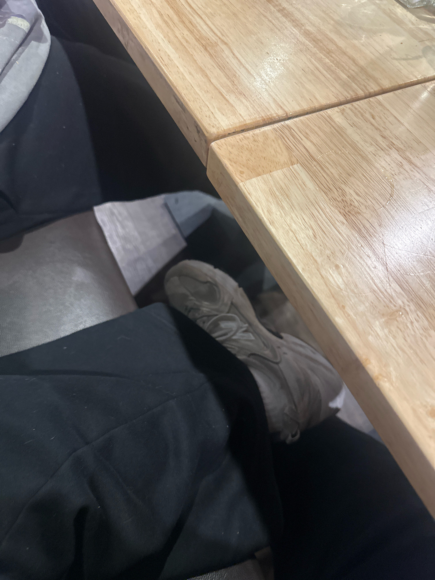 A person's foot wearing a light brown athletic shoe is visible beneath a light wood table. The person is wearing black pants. The environment appears to be indoors, possibly a cafe or restaurant, with a dark upholstered seat visible. There is no indication that this image is located in Salisbury, Australia.