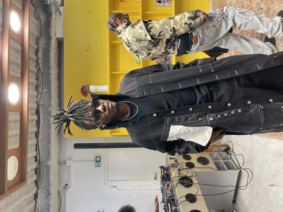 This indoor scene depicts three individuals within a commercial or event space, consistent with venues found in Paris, France. The primary subject, a dark-skinned male, stands center-left, angled slightly right of the viewer. He wears dark sunglasses and has dark dreadlocks styled into an upright top bun. His attire consists of a long, dark, button-front coat with numerous silver snaps, featuring a large, distressed white fabric patch on the left side, worn over a black t-shirt and dark pants. A thin silver chain adorns his neck, and his right hand is partially obscured, possibly in a pocket. To his immediate left, partially visible, a dark-skinned individual operates a DJ setup on a light-colored console with multiple circular cutouts and exposed cables. The setup includes two Pioneer CDJ units and a Pioneer mixer. A tall, black speaker column stands behind the primary subject. To the right, a second dark-skinned male is viewed from behind, dressed in a camouflage jacket, heavily distressed light-wash jeans with extensive fraying, and a black fanny pack bearing "Supreme" branding across the lower back. The background features a bright yellow shelving unit displaying various items, including a mannequin head wearing a red, white, and blue striped knitted cap. The ceiling is light-colored and textured, with exposed conduits and circular recessed lights framed by wood. The floor is tiled, with a patterned rug visible beneath the feet of the person on the right. A white wall on the far left shows visible cracking and displays a green ISO 7010-compliant exit sign (running figure, white arrow) with a red fire extinguisher symbol below it, which is standard safety signage used in Paris and across Europe.