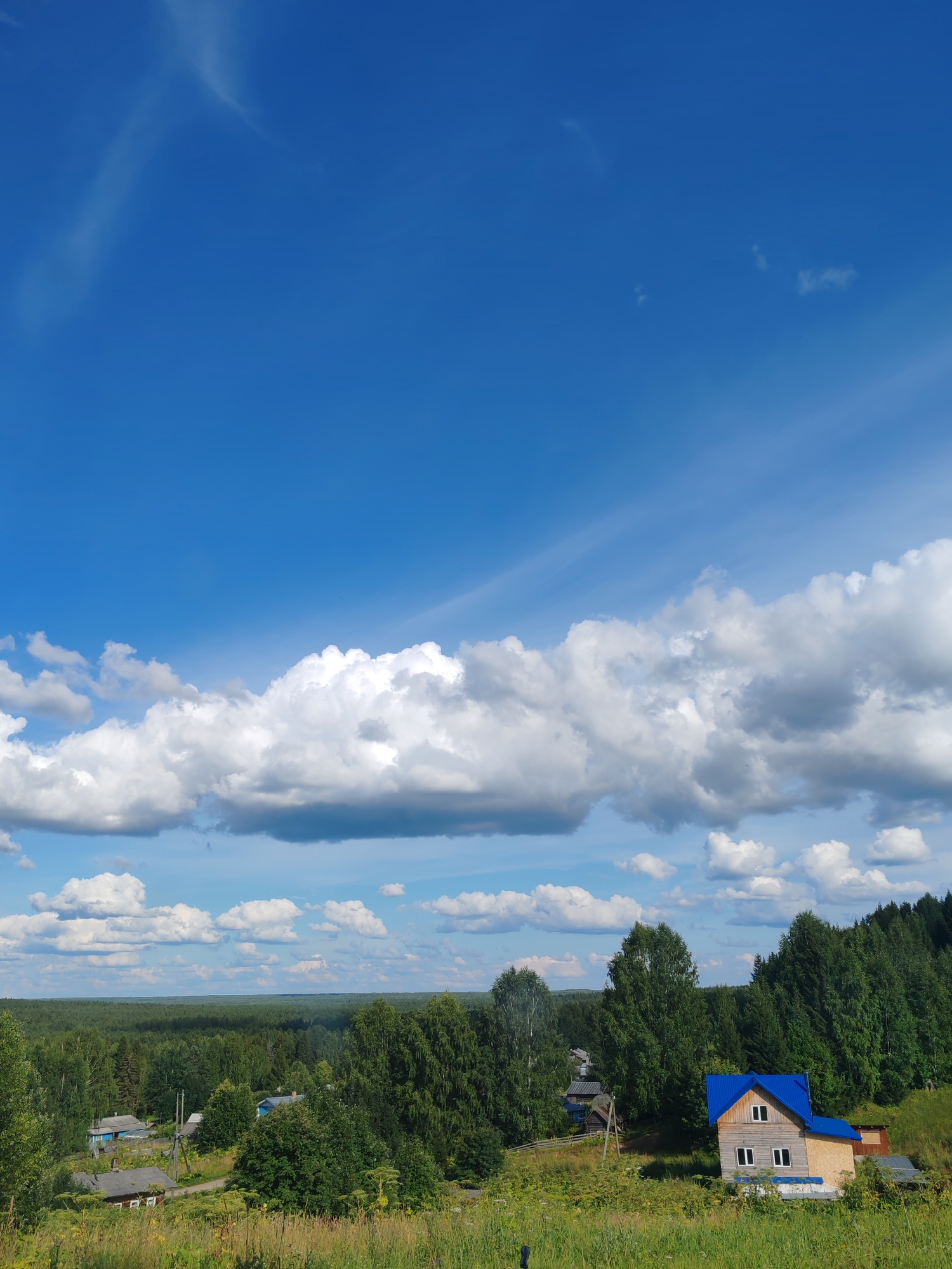 The image depicts a rural landscape under a bright blue sky with scattered white clouds. In the foreground, a grassy hill slopes upwards. A small village is nestled among lush green trees. Several houses are visible, some with blue roofs, and a larger house with a prominent blue roof is situated on the right side of the image. A dense forest borders the village in the background. The scene is tranquil and appears to be taken on a sunny day. The location is identified as Chukhlem, Russia.