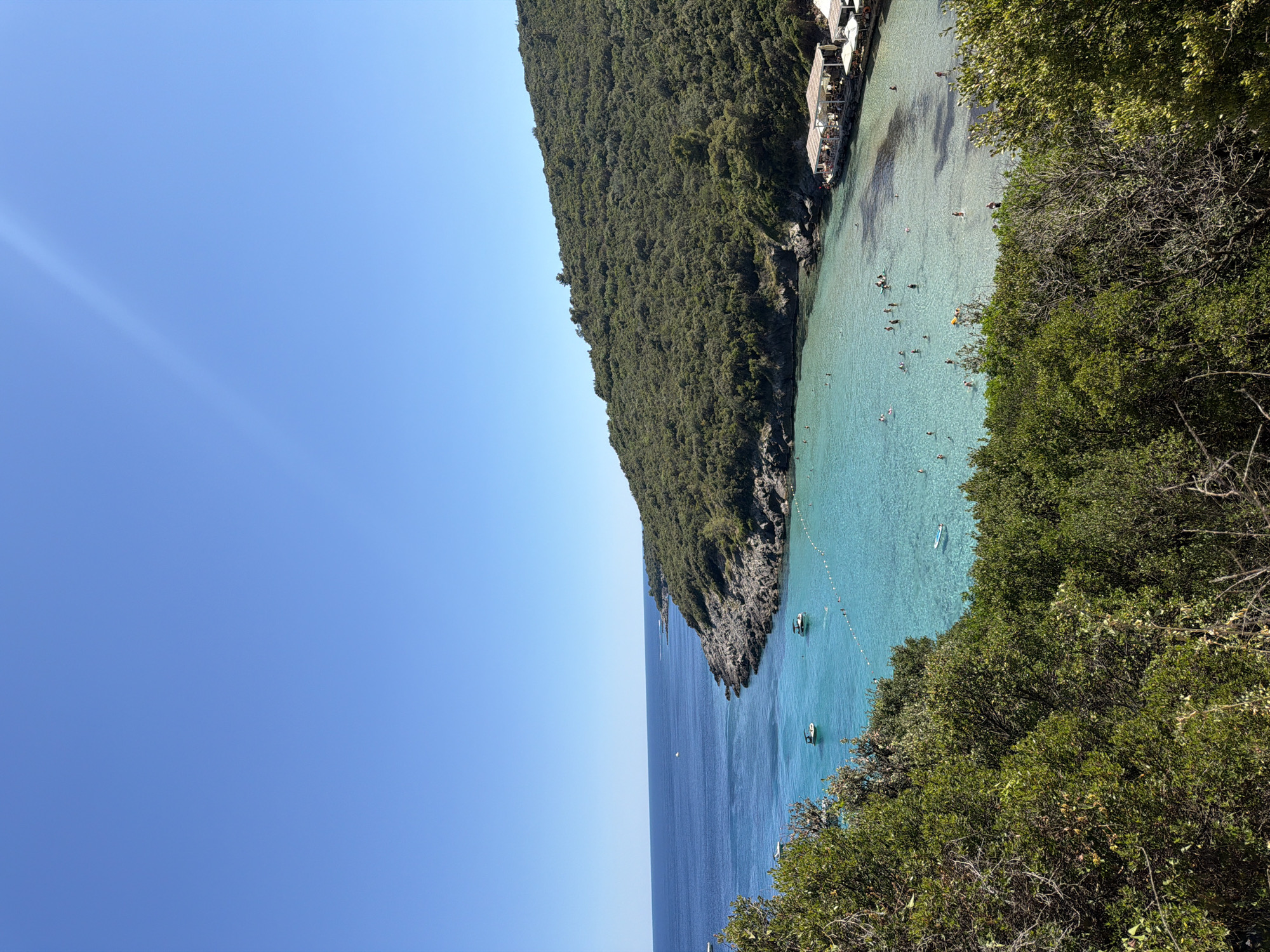 This image captures a scenic view of a cove in Prijevor, Montenegro, under a clear blue sky. The foreground is dominated by lush green foliage, with trees and shrubs obscuring part of the view. Beyond the vegetation, a vibrant turquoise bay unfolds, dotted with several small boats. The water is exceptionally clear, revealing the sandy bottom in shallower areas. A small beach is visible along the edge of the bay, where a cluster of white-roofed structures, likely beach bars or restaurants, are situated. Numerous people are present in the water, swimming and enjoying the sea. Rocky outcrops and a densely vegetated hillside form the backdrop of the cove, leading to the open sea.